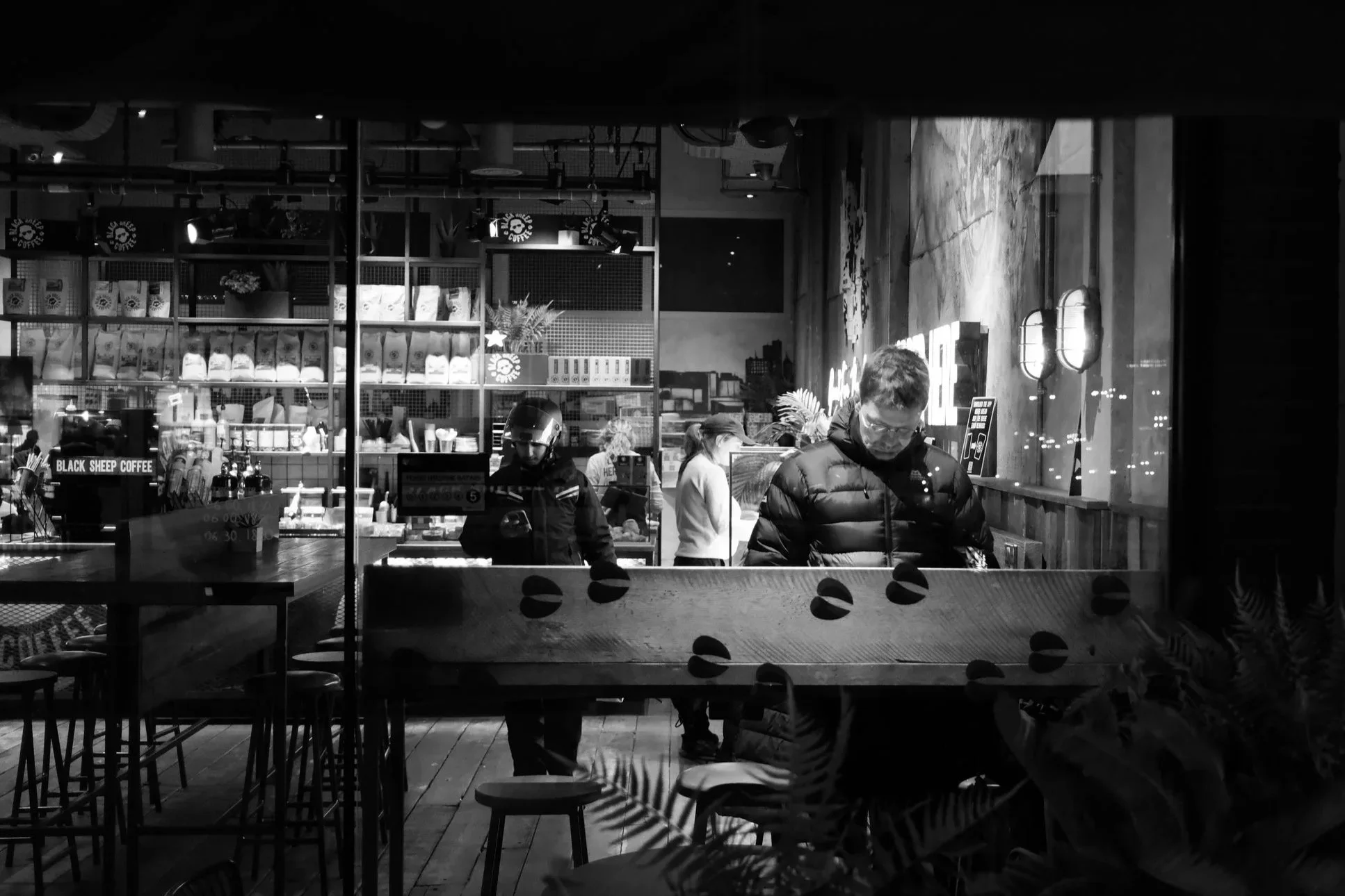Inside a modern coffee shop at night with customers and staff, shelves of coffee supplies, and warm lighting.