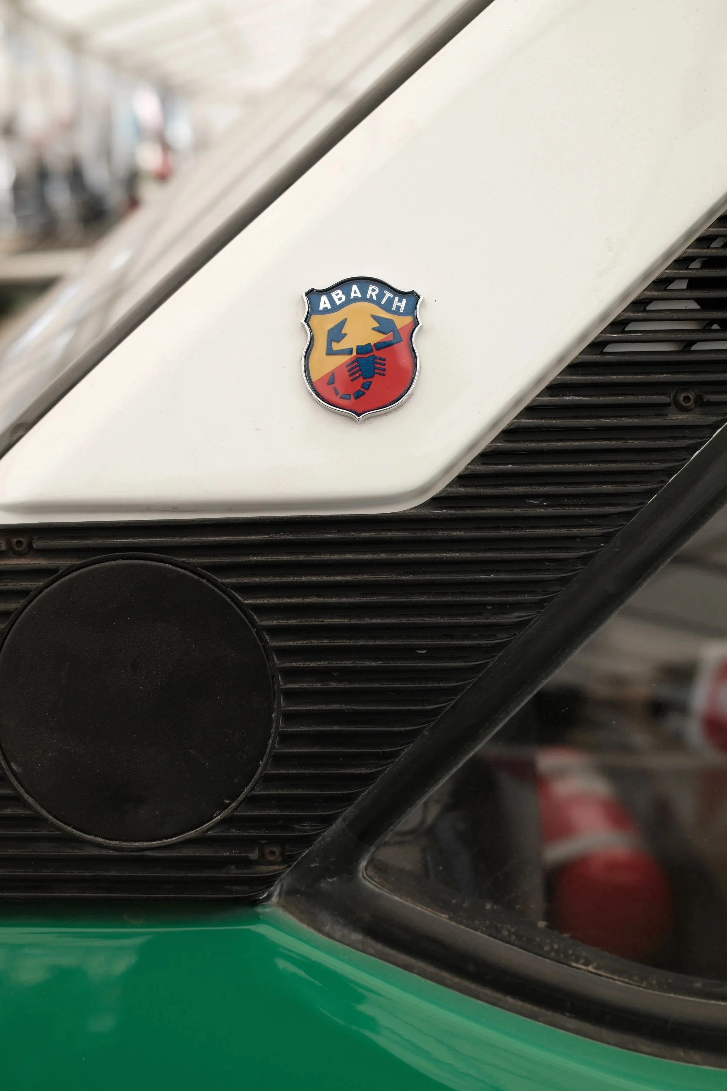 Close-up of a white vehicle with an Abarth badge on the front. The badge features a shield with a red, yellow, and black scorpion logo and the word "ABARTH" at the top. The vehicle is parked near a green surface.