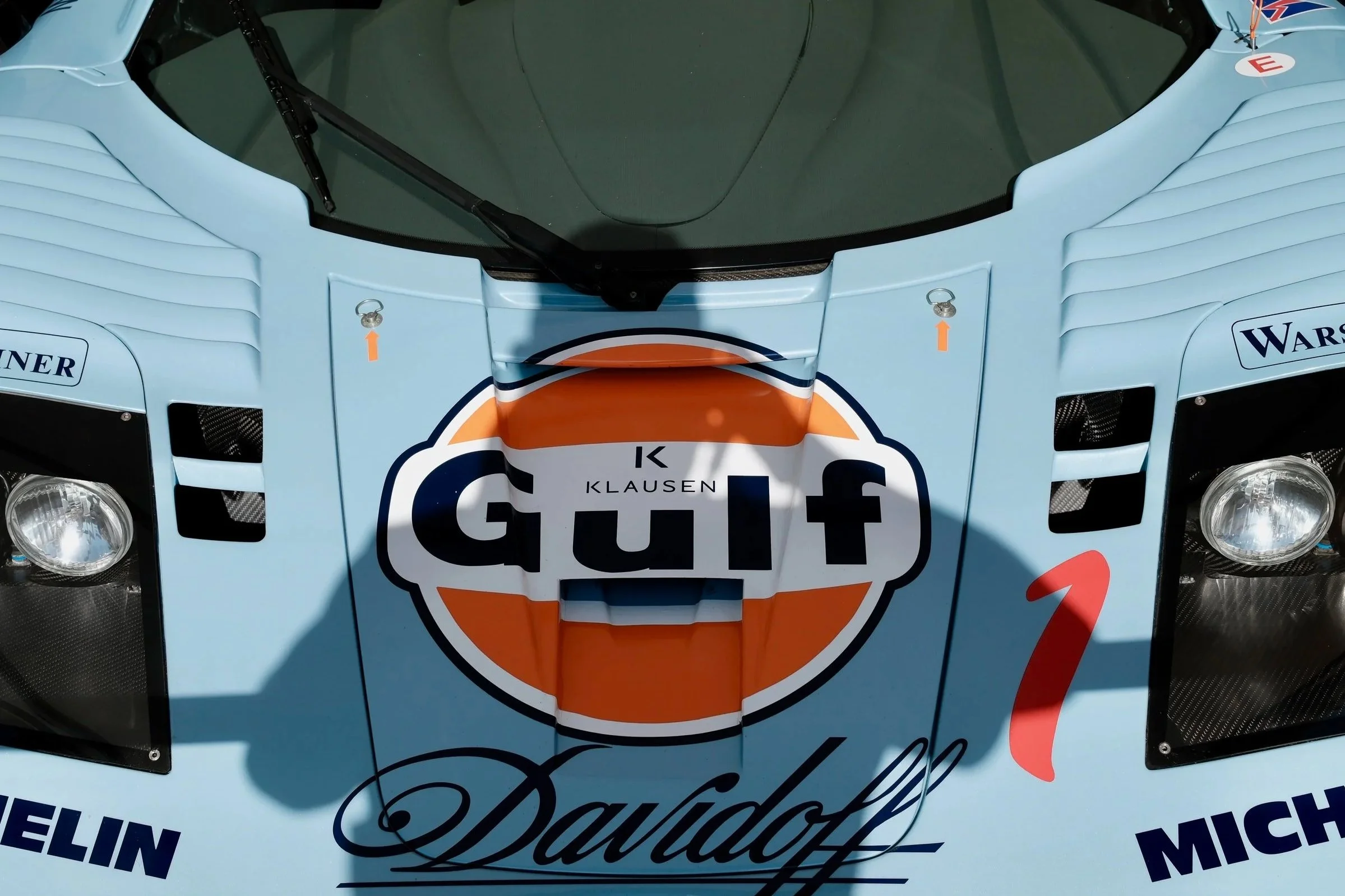 Close-up view of a race car's front, featuring various sponsor stickers, a large logo reading 'Dauphin,' and two aerodynamic mirrors attached to the hood.