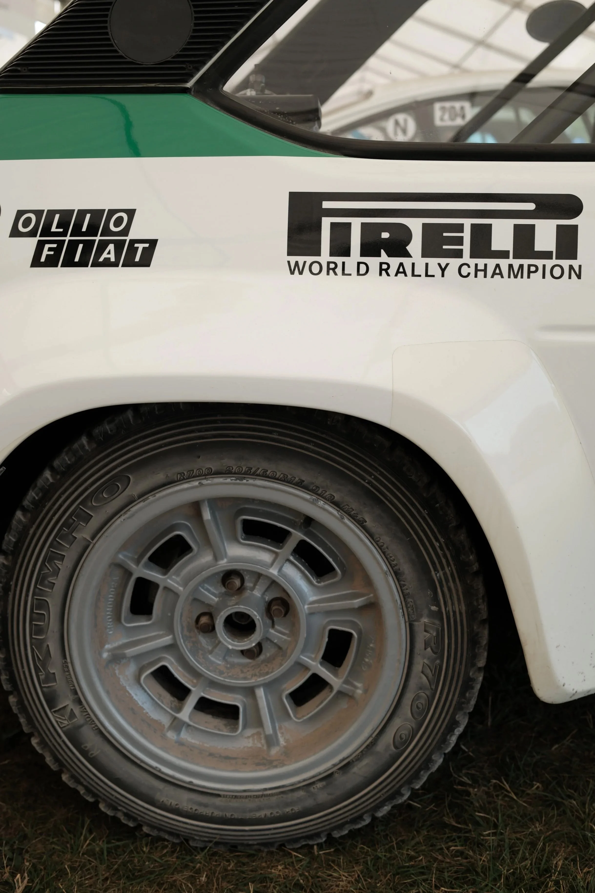 Close-up of a vintage white rally car's front fender with Diatto and Pirelli logos, showing a silver alloy wheel with a rugged tire on grass.