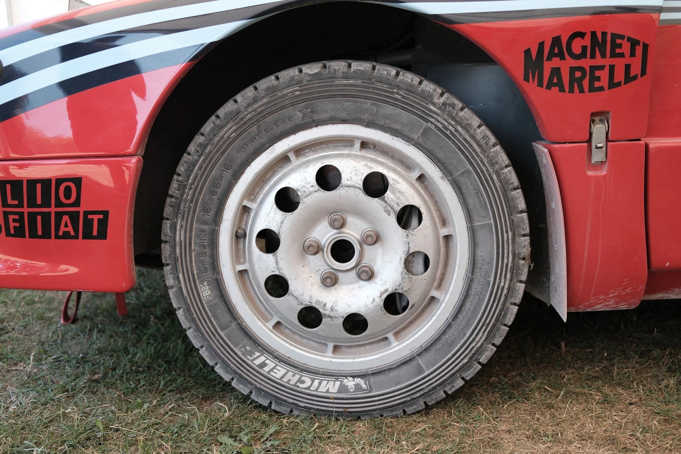 Close-up of a race car wheel and tire mounted on a red and black race car, with sponsor logos and stickers including "MAGNETI MARELLI" and a logo with the letters "LIOFIAT." The car is on grass.