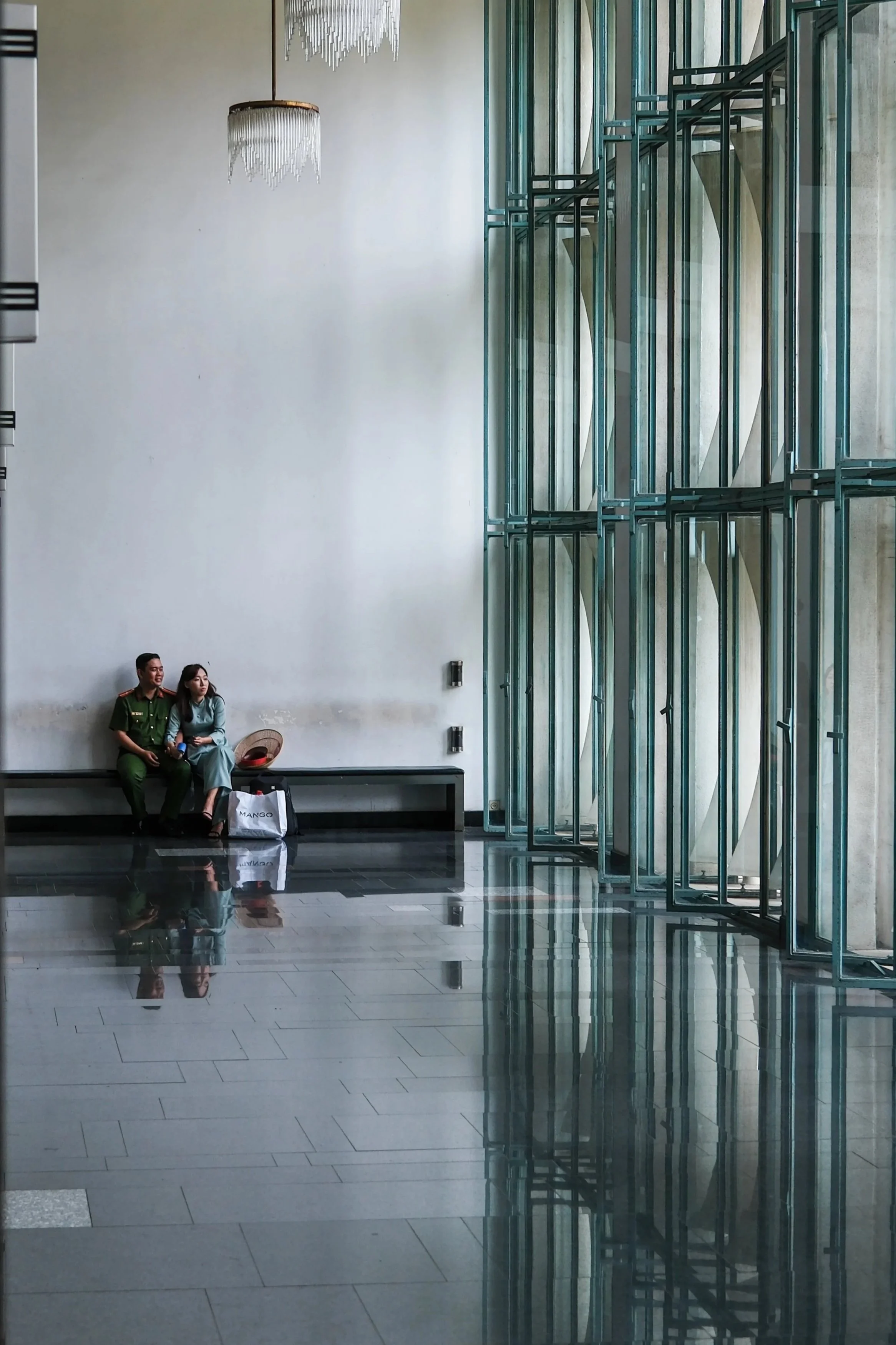 Two people sitting on a bench in a modern building with large windows and reflective tile flooring. One person is in a military uniform, and the other is in casual clothing. There are shopping bags placed beside them, and two chandeliers hanging from