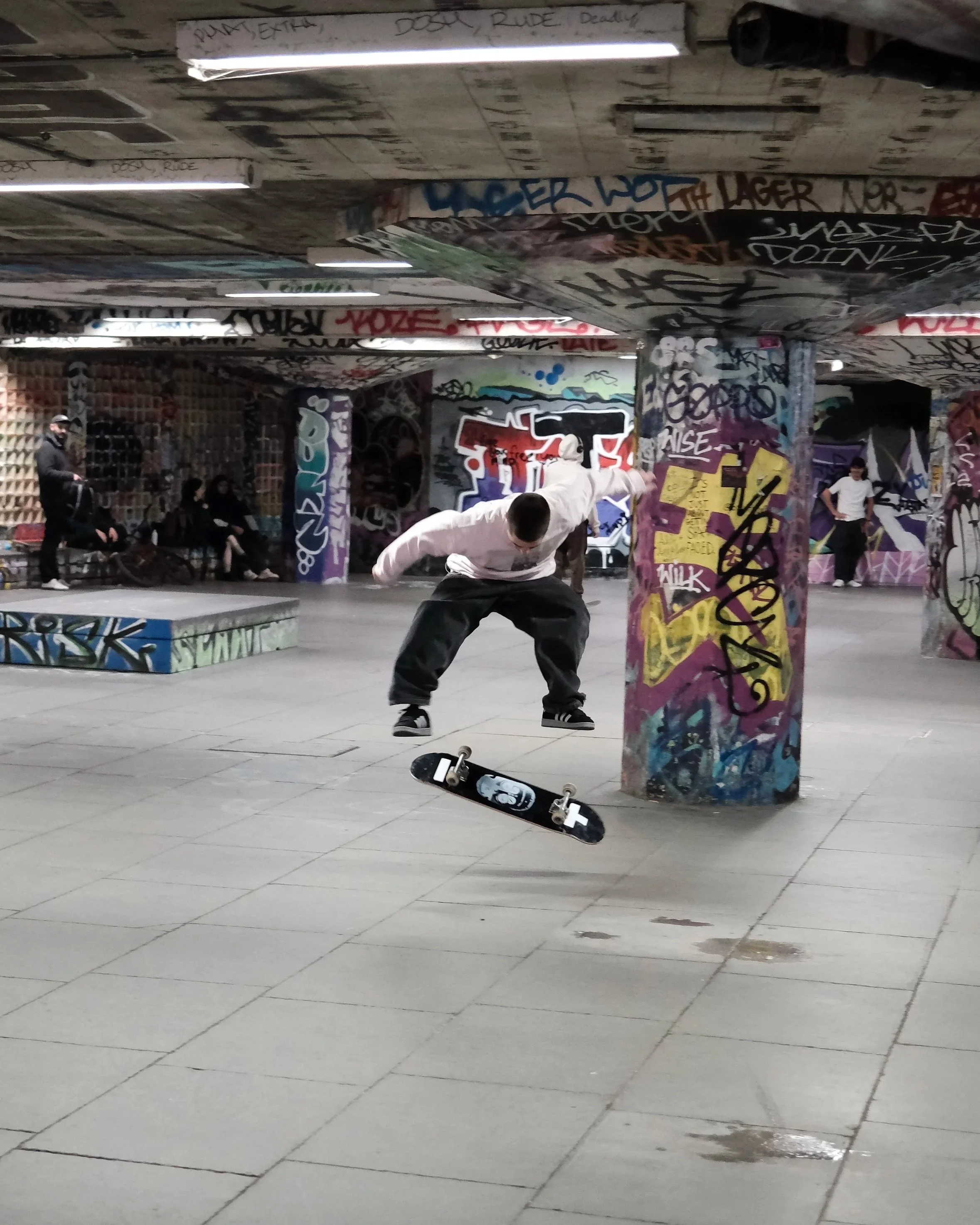 A young man does a skateboard trick in an underground skate park with graffiti-covered pillars and walls. Other people are sitting and standing nearby, with graffiti tags and messages on the walls and ceilings.