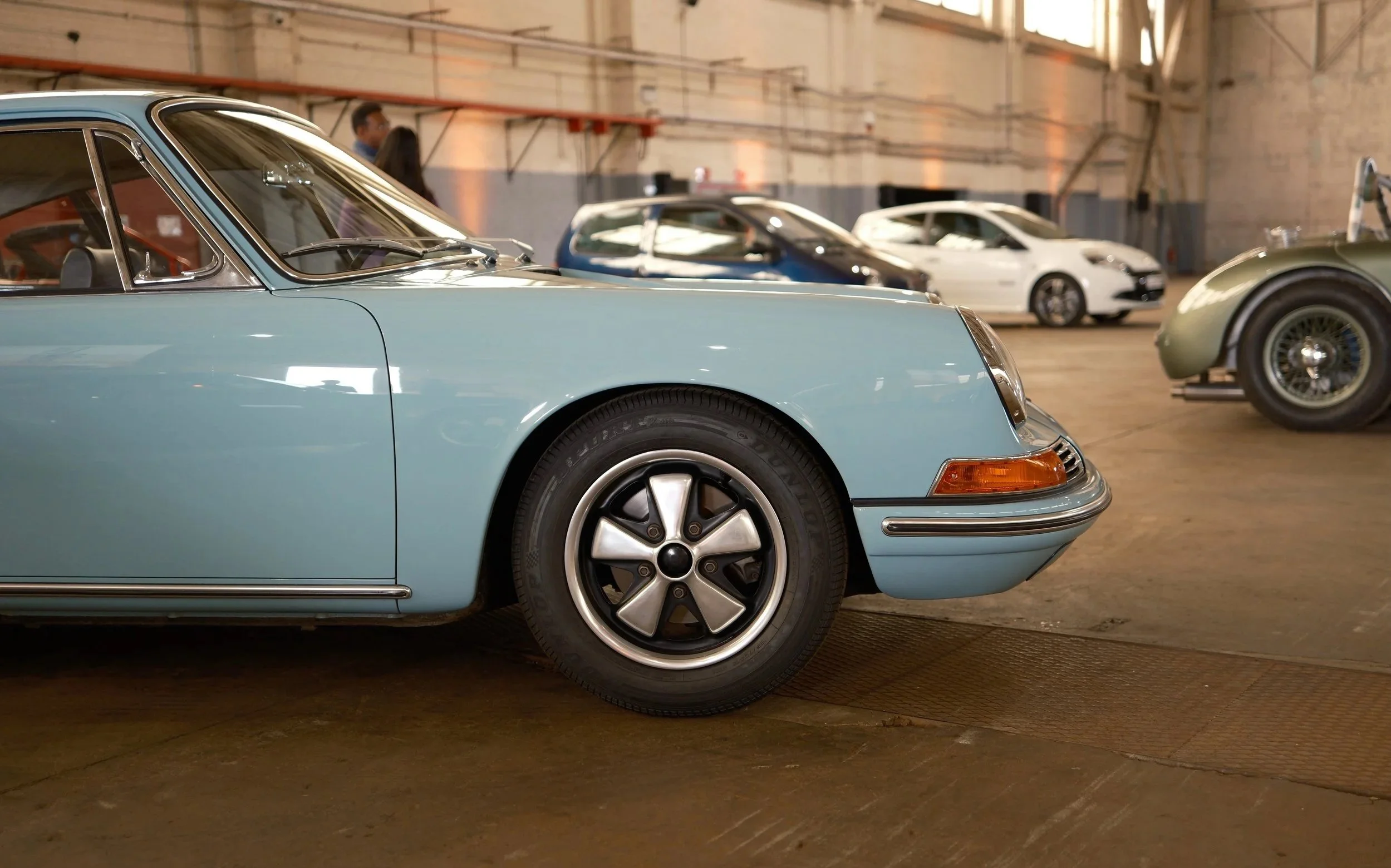 Close-up of a vintage light blue car with sleek design, parked indoors in a warehouse with other modern cars in the background.