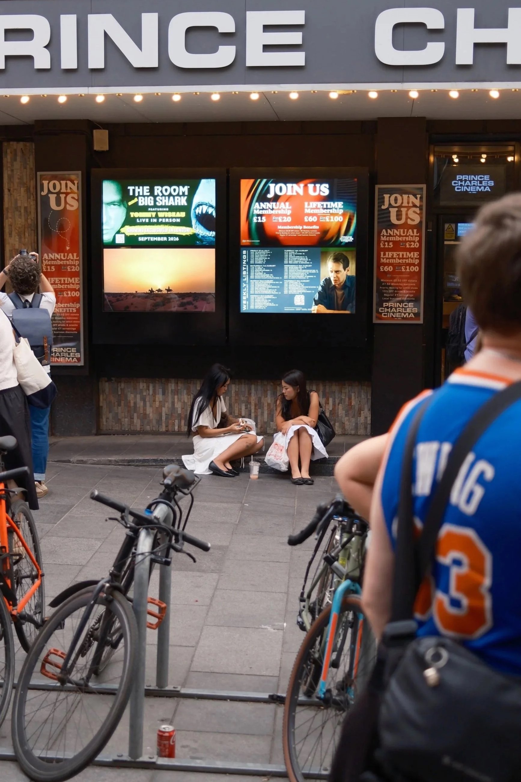 People standing and sitting outside the Prince Charles Cinema, with bicycles parked in front. Digital screens display information about movies and memberships.