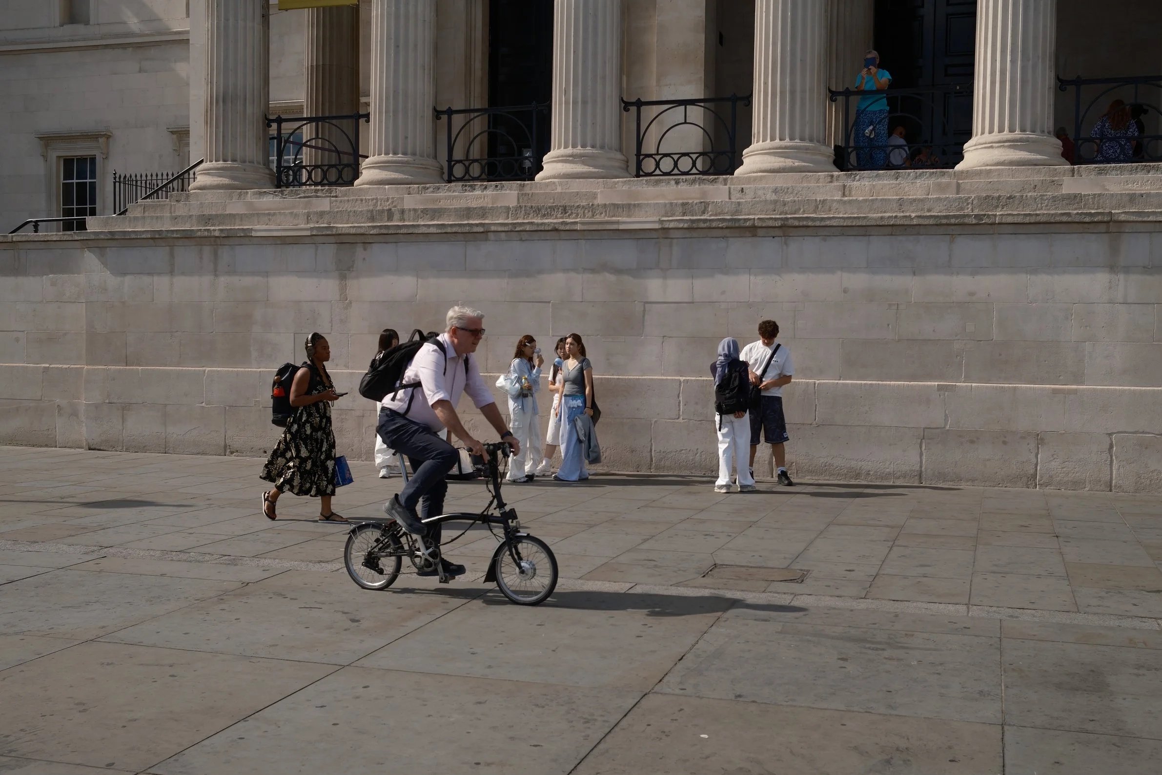 People standing and walking in front of a large building with classical columns, including a man riding a bike in the foreground and several women and men on the steps or sidewalk.