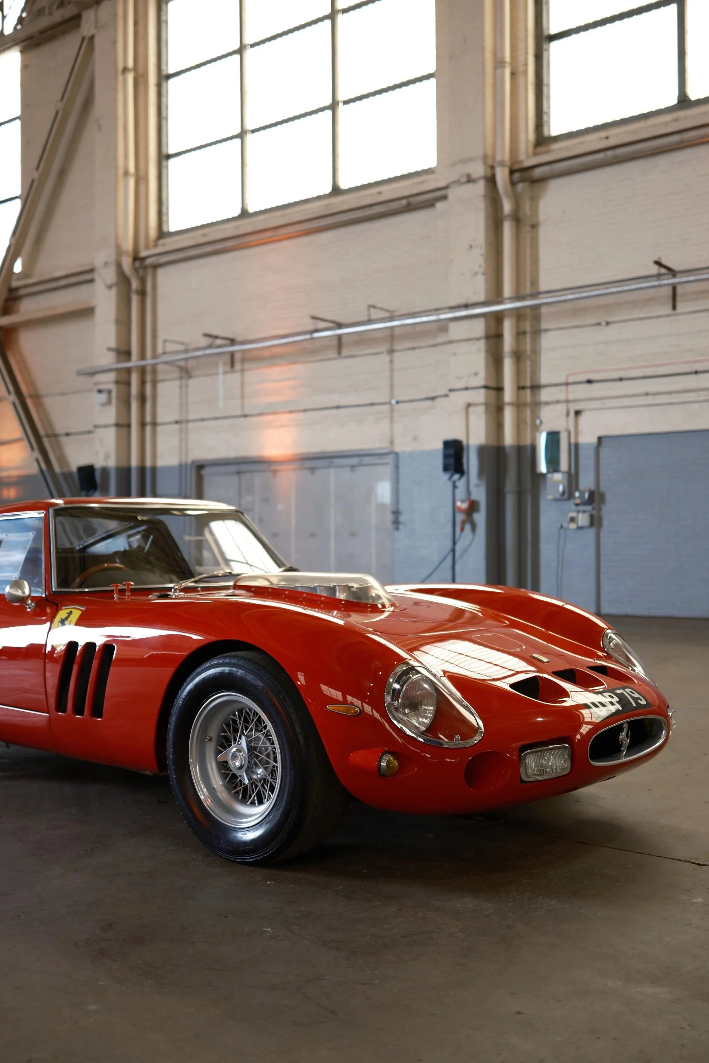 A red vintage race car with a sleek, aerodynamic design parked inside an industrial-style warehouse with high windows and exposed pipes.