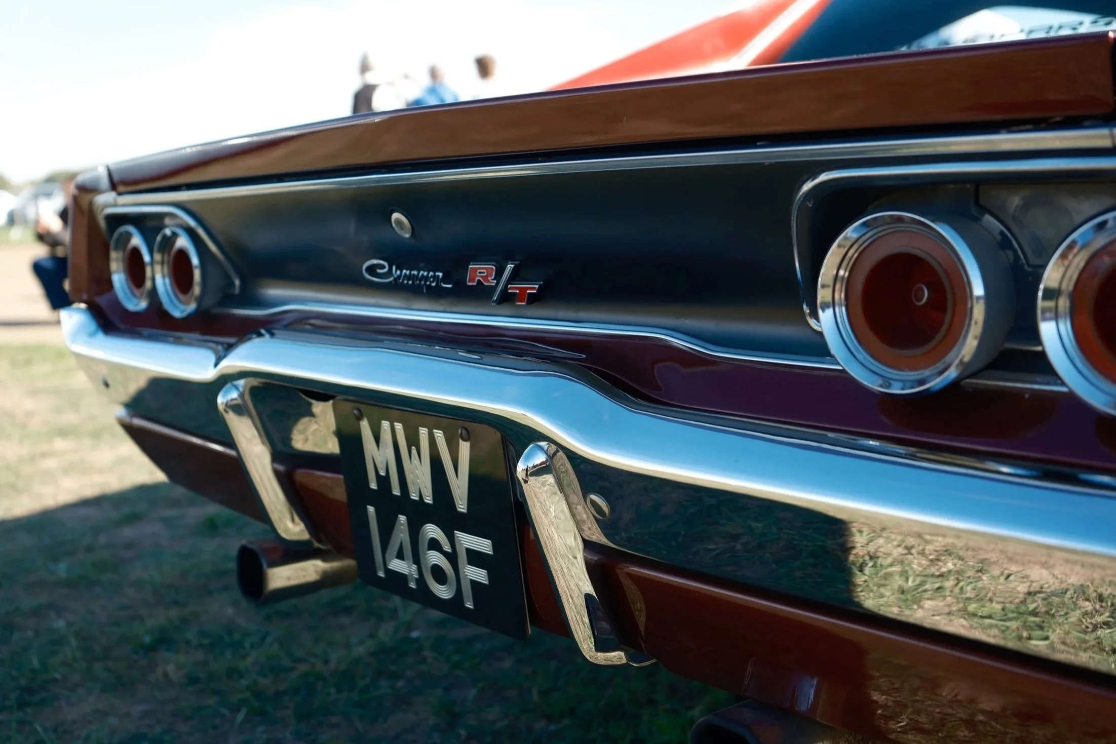 Close-up of the rear end of a vintage black muscle car with dual exhaust pipes and a wooden rear spoiler at an outdoor car show, with people in the background.