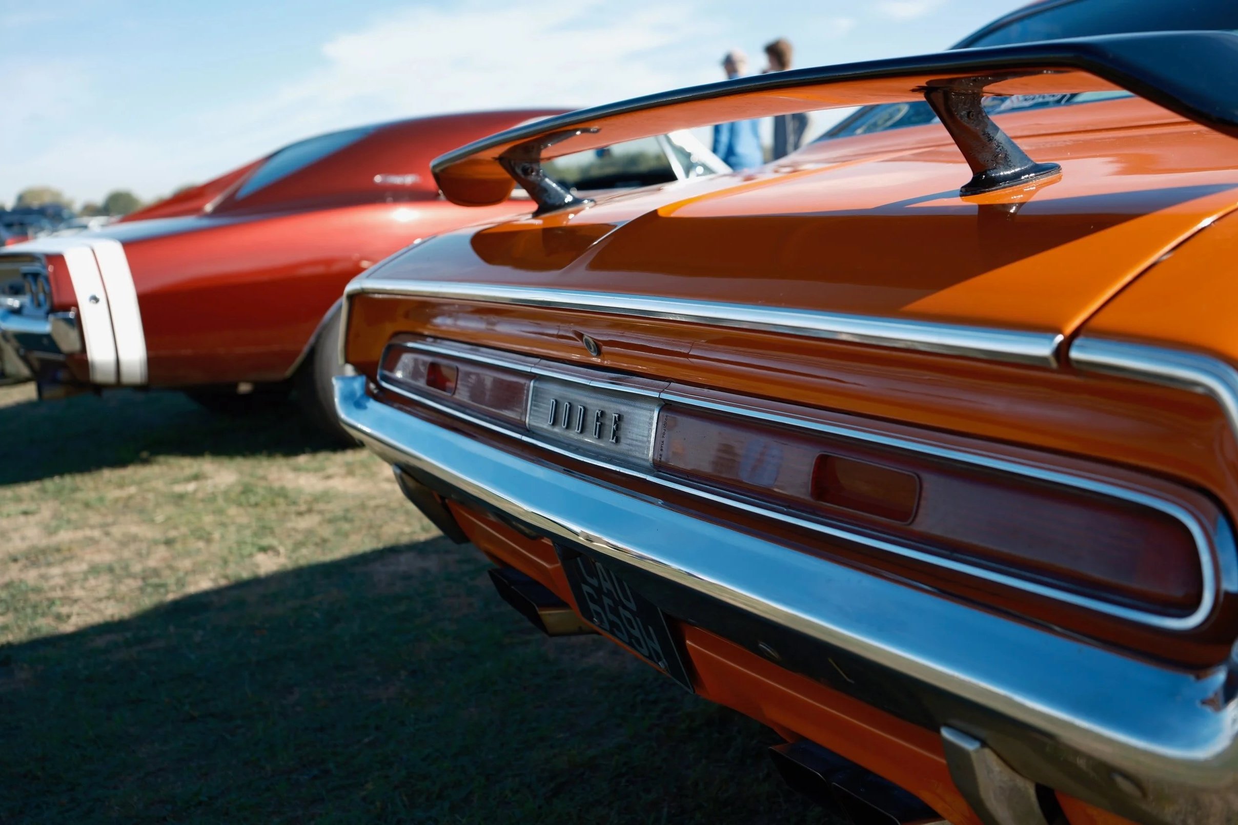 Close-up of the rear of an orange classic Dodge muscle car with a prominent rear spoiler, parked outdoors on grass, with other cars and people blurred in the background.