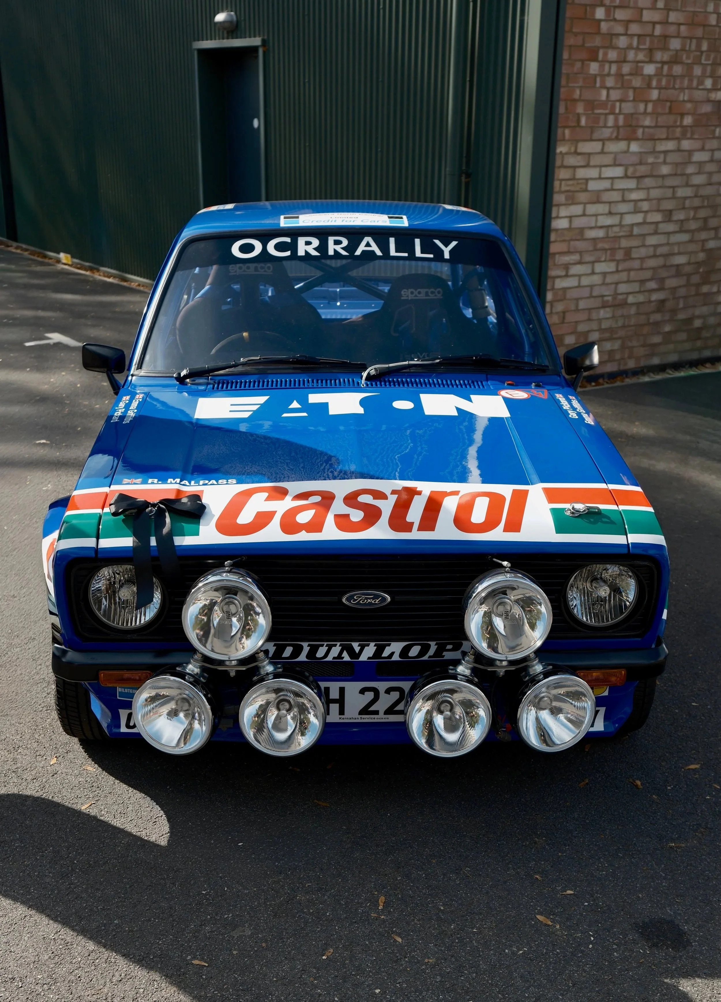 Front view of a blue vintage rally car with four large auxiliary lights mounted on the bumper, branded with Castrol and Dunlop logos, parked outdoors near a brick wall and a metal building.