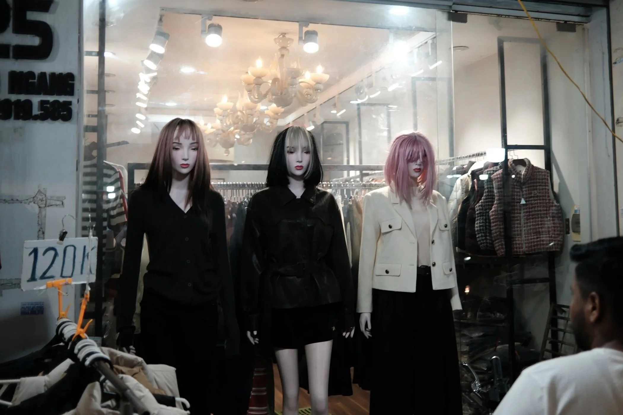 A storefront window display shows three mannequins with different hairstyles and outfits, inside a clothing store with chandeliers and racks of clothing visible in the background.