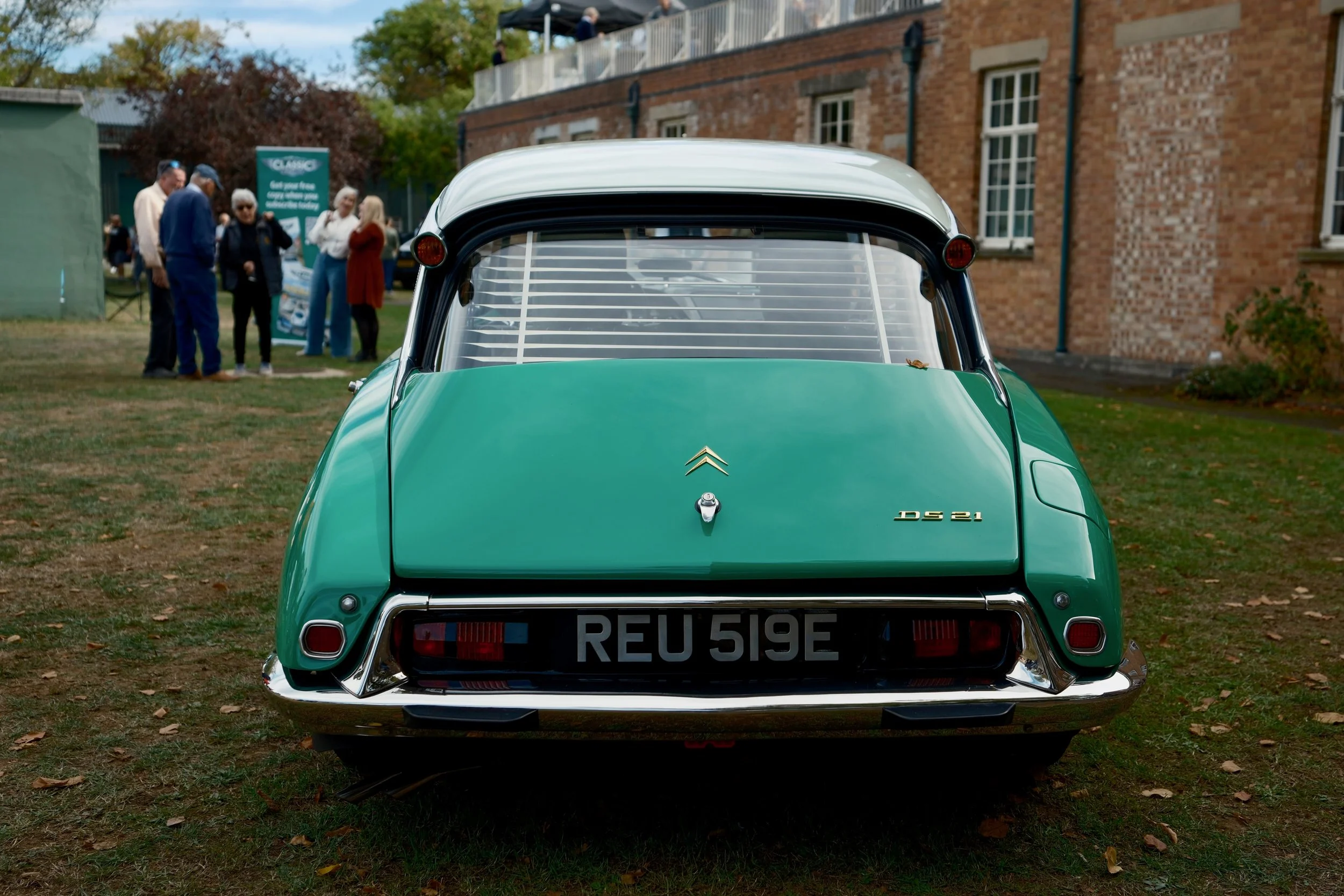 Rear view of a vintage green Citroën DS 21 car parked on grass at an outdoor event, with a group of people and brick building in the background.