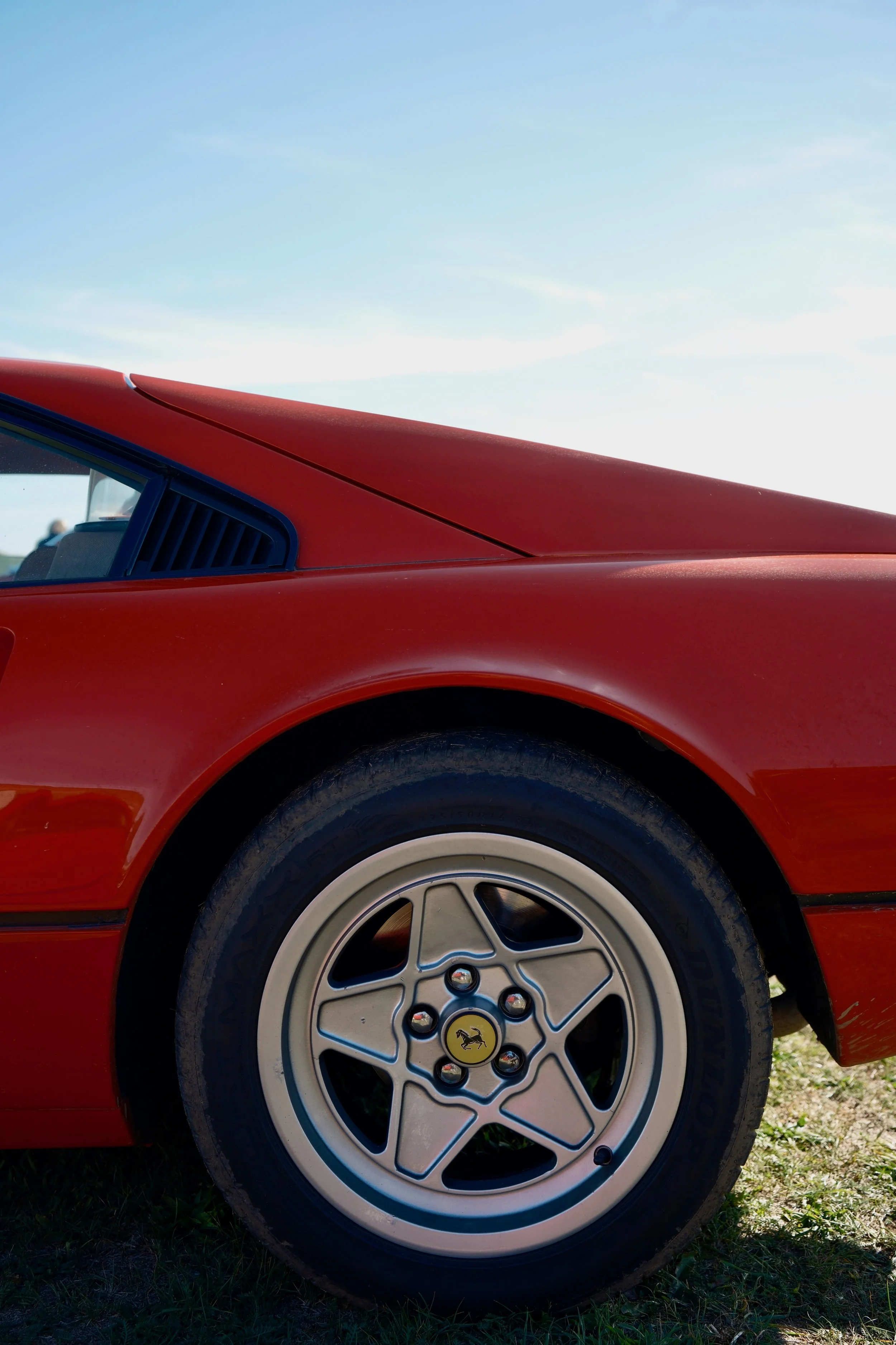 Close-up of the front wheel and side of a red vintage Ferrari car on grass under a blue sky.