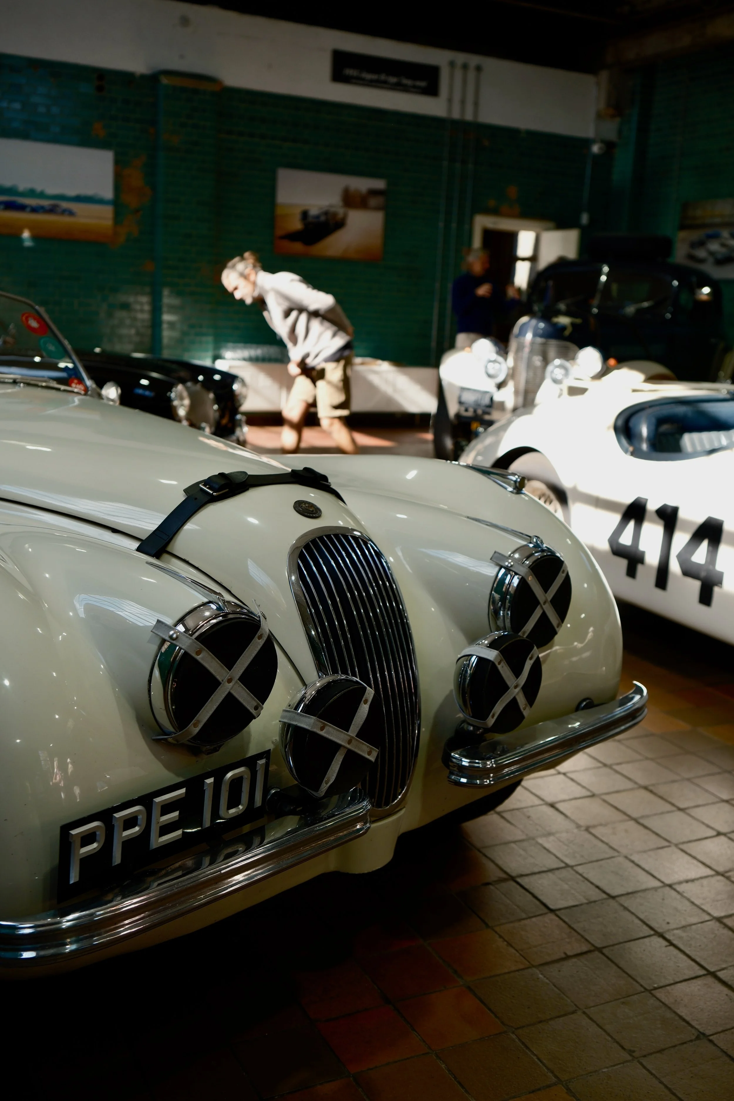 A vintage white sports car with a black strap across the hood, displaying the license plate 'PE101L' and classic front grille and headlights. In the background, there are other classic cars and a person walking while looking down, inside a museum or 