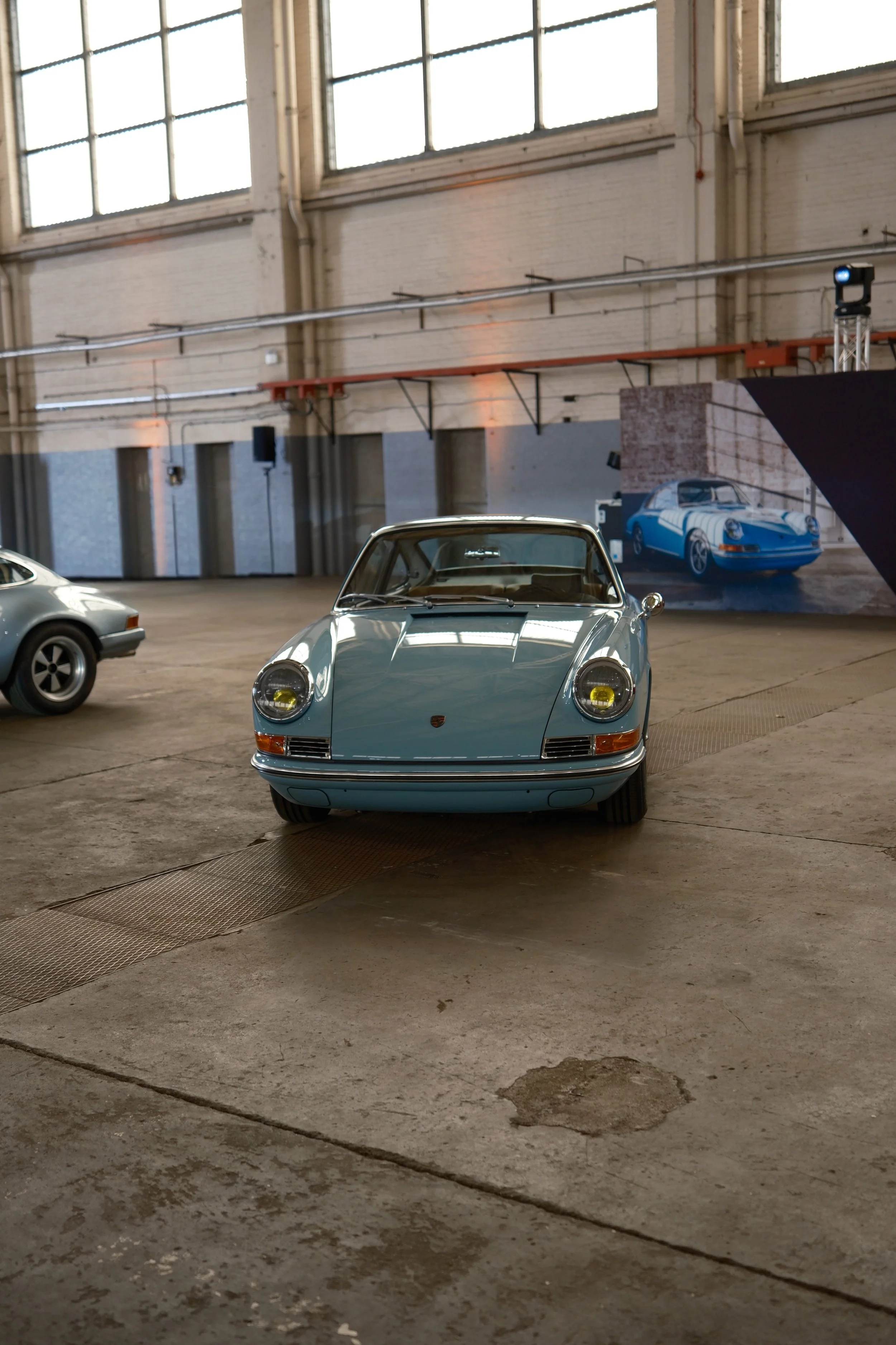 A vintage light blue Porsche 911 sports car is in the foreground of an indoor space with other cars and a large mural of another Porsche in the background.