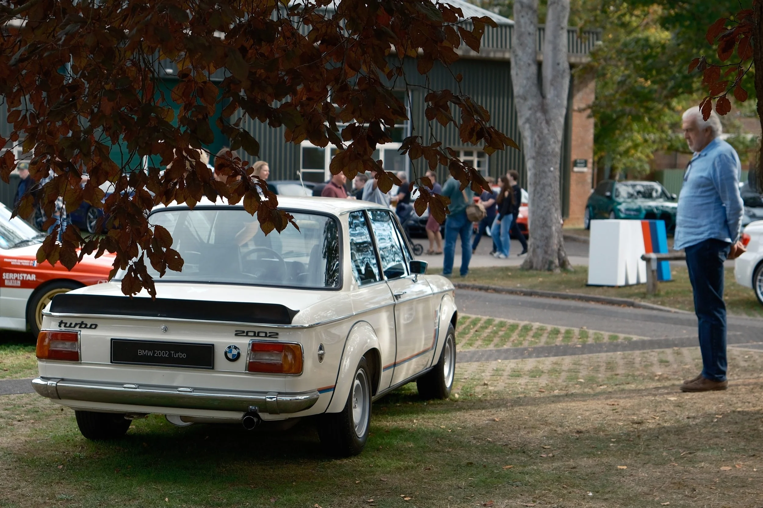 A white vintage BMW 2002 Turbo parked on grass with a black rear bumper, set amidst an outdoor display of classic cars and people gathered around, with trees and a building in the background.