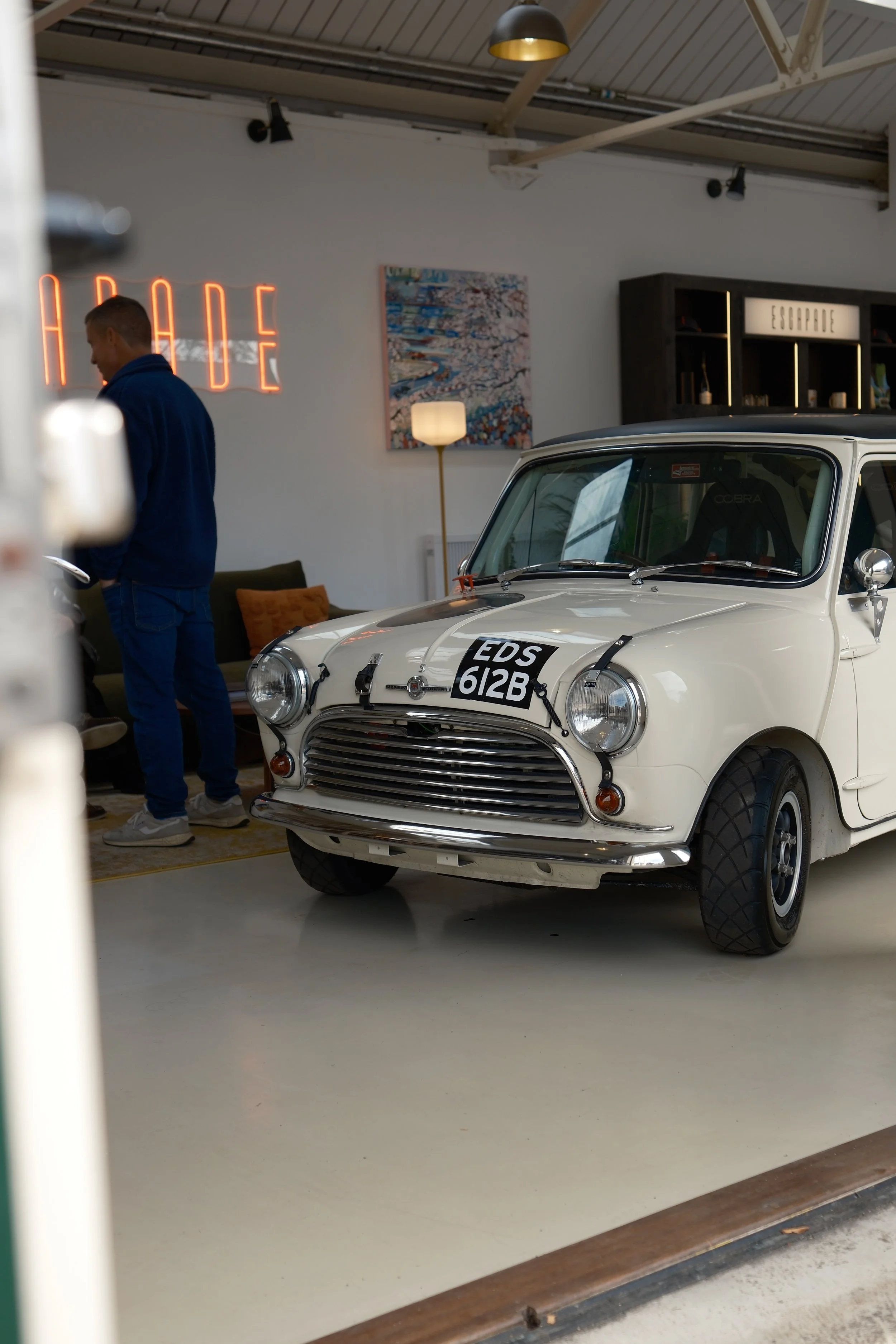 A white miniature car with black tires parked inside an industrial-style room, with a man in a blue hoodie standing nearby and a red neon sign on the wall that reads "HEROES." The room has white walls, a colorful abstract painting, a floor lamp, and 