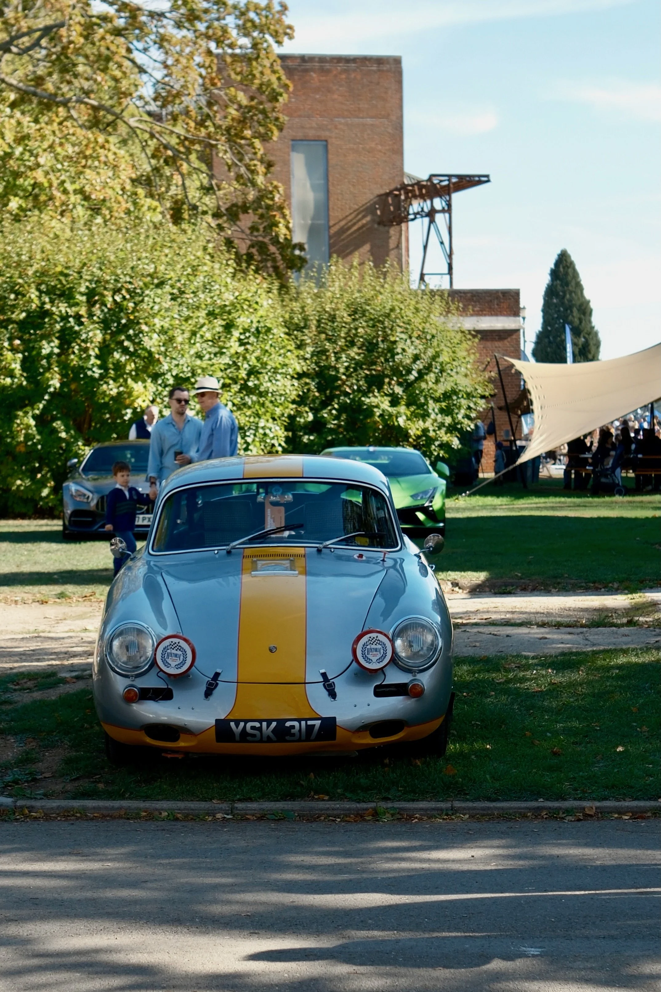 A silver vintage race car with yellow stripe, rally stickers, and a British license plate YSK 317 parked in front of a grassy area with trees and a brick building, possibly during a car show.