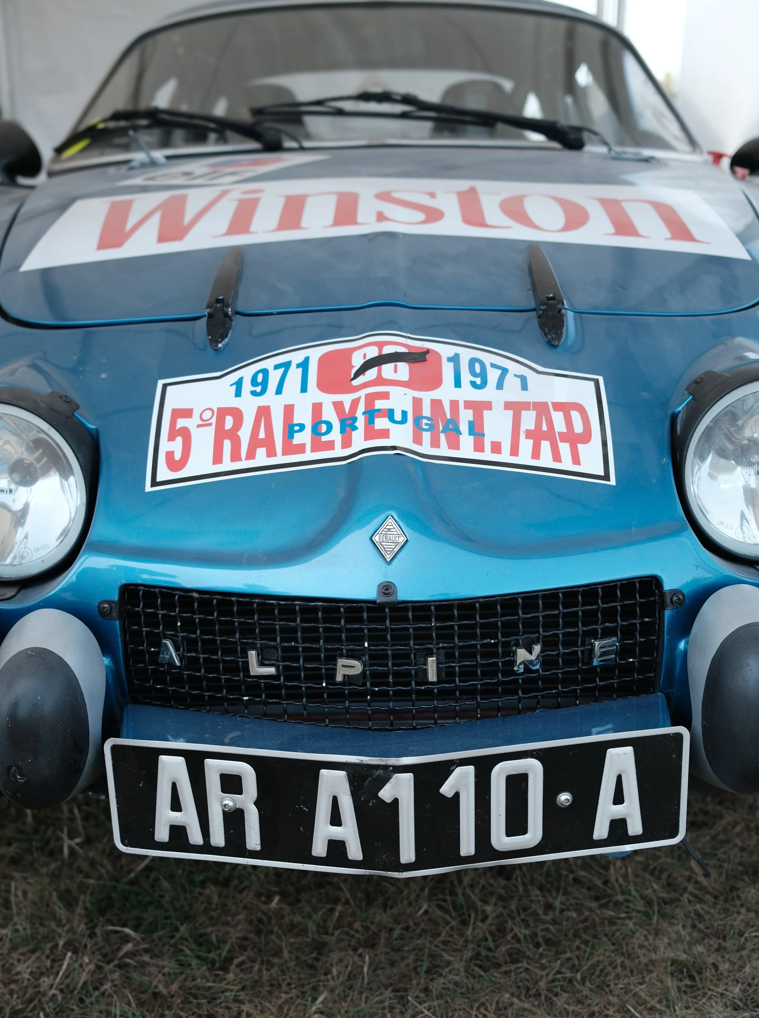 Front view of a vintage blue race car with rally stickers, rally number, and a license plate, parked on grass.