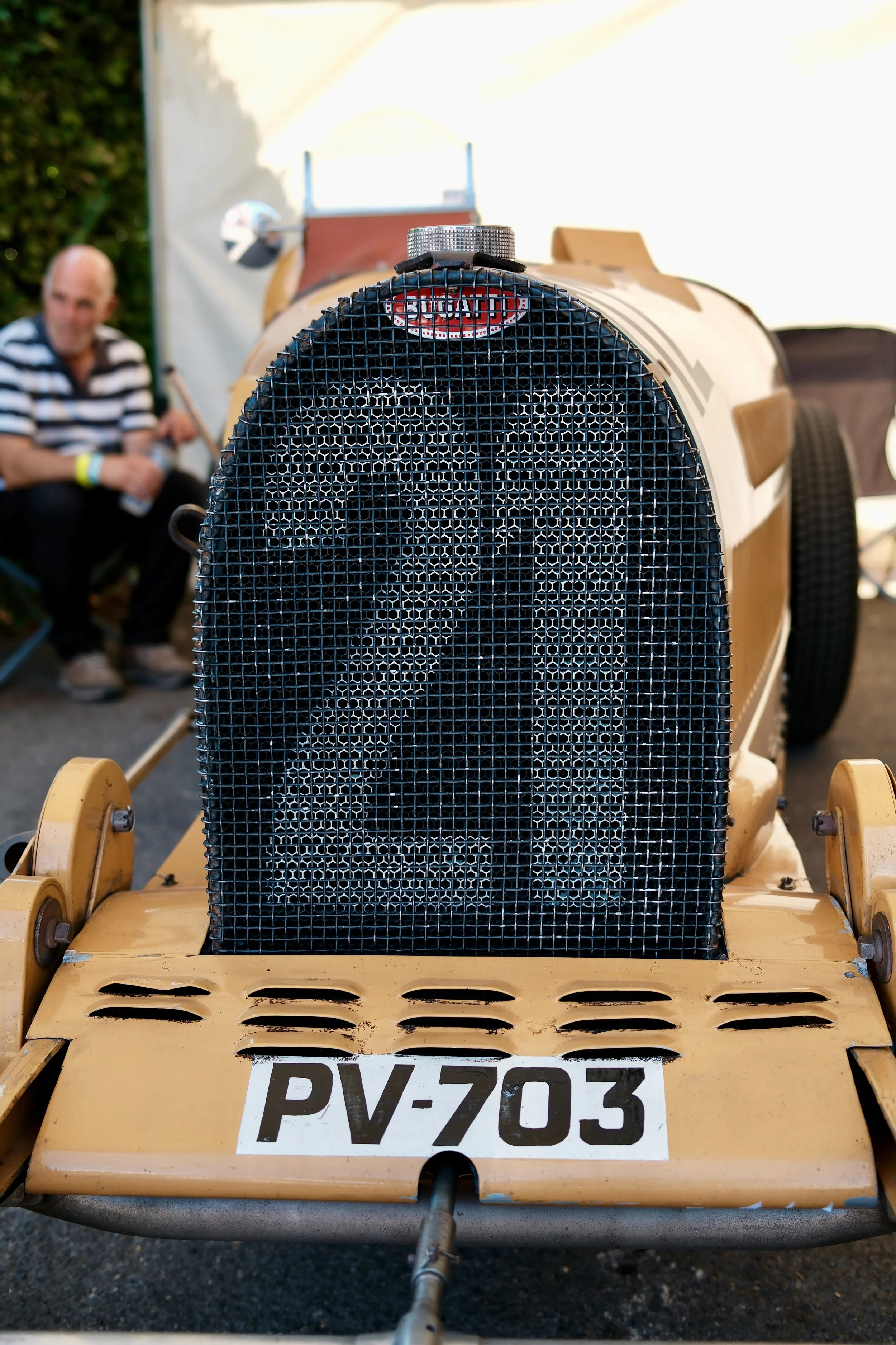 Close-up of the front grill of a vintage yellow go-kart with the license plate PV-703, and a man in a striped shirt sitting in the background.