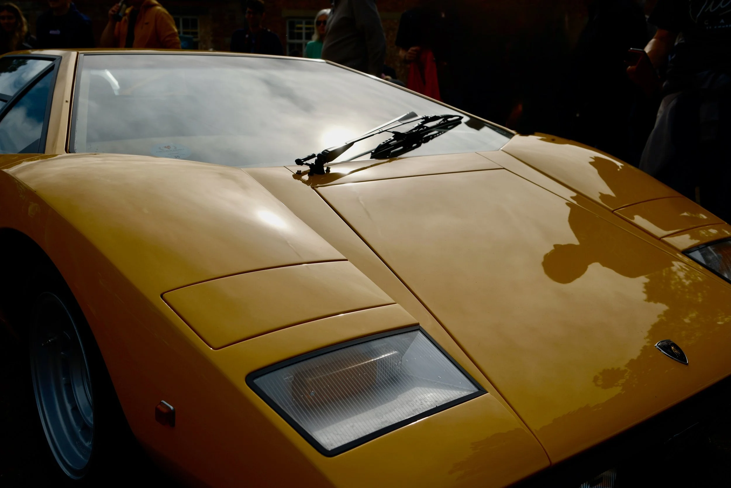 A close-up of a yellow vintage sports car with a sleek design, parked outdoors during daytime with people in the background.
