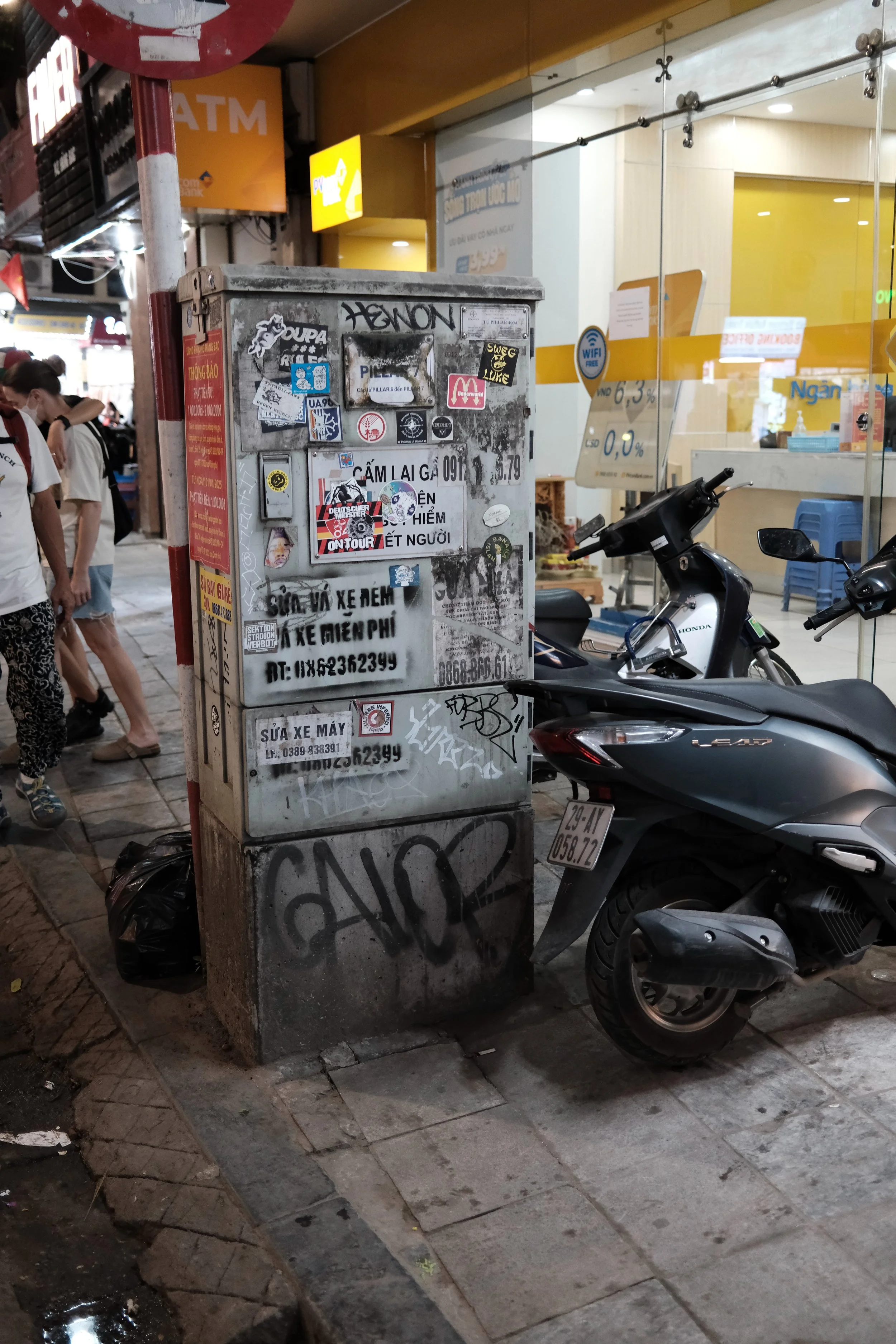 A street scene at night showing a bulletin board with various stickers and advertisements, a parked motorcycle, a shop with a yellow sign, and several people walking by.
