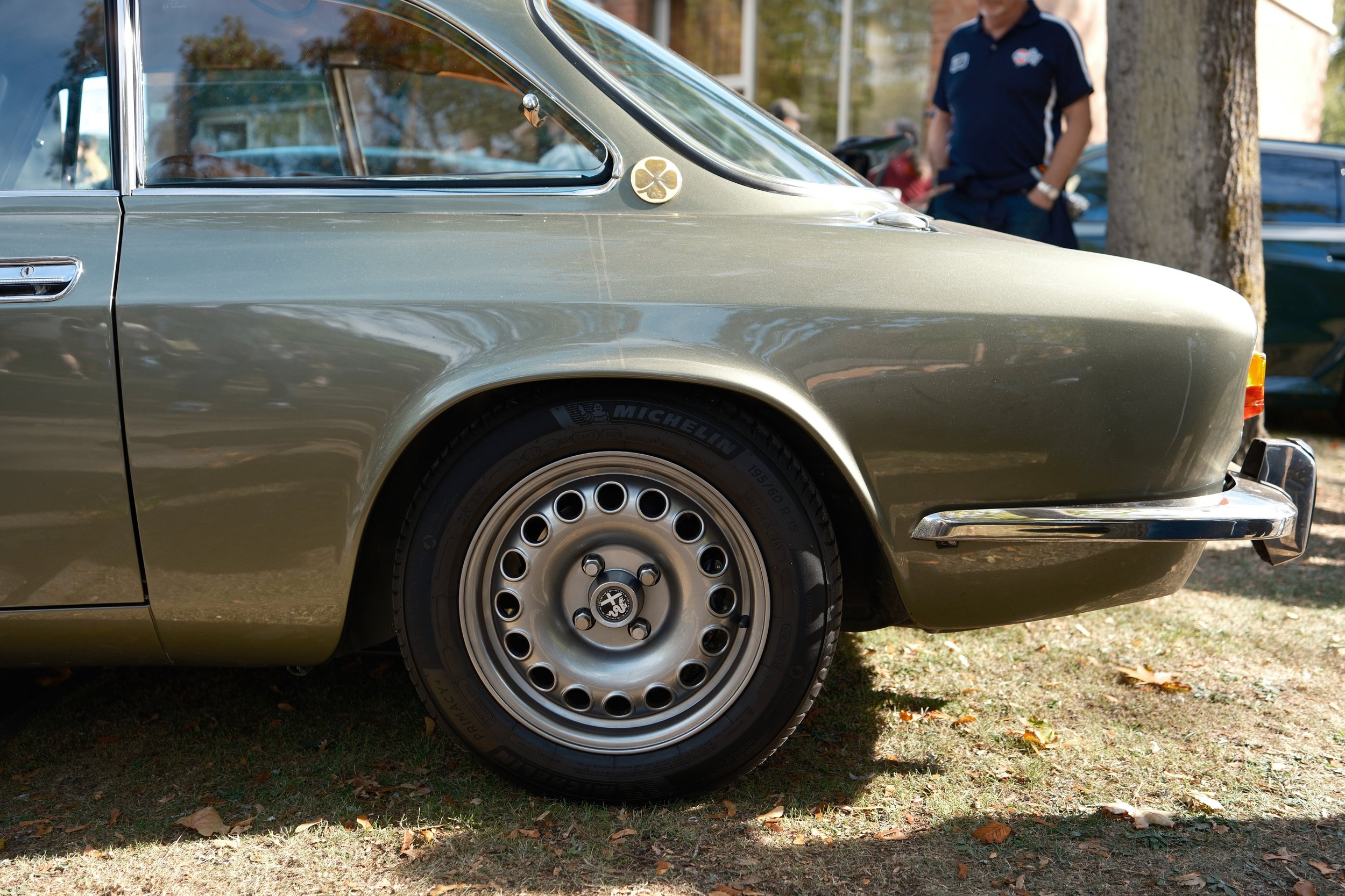 Close-up of the side of a vintage silver-gray car parked on grass with fallen leaves, showing the wheel, door, window, and a small emblem on the fender, with a man in a dark sports shirt standing nearby in the background.