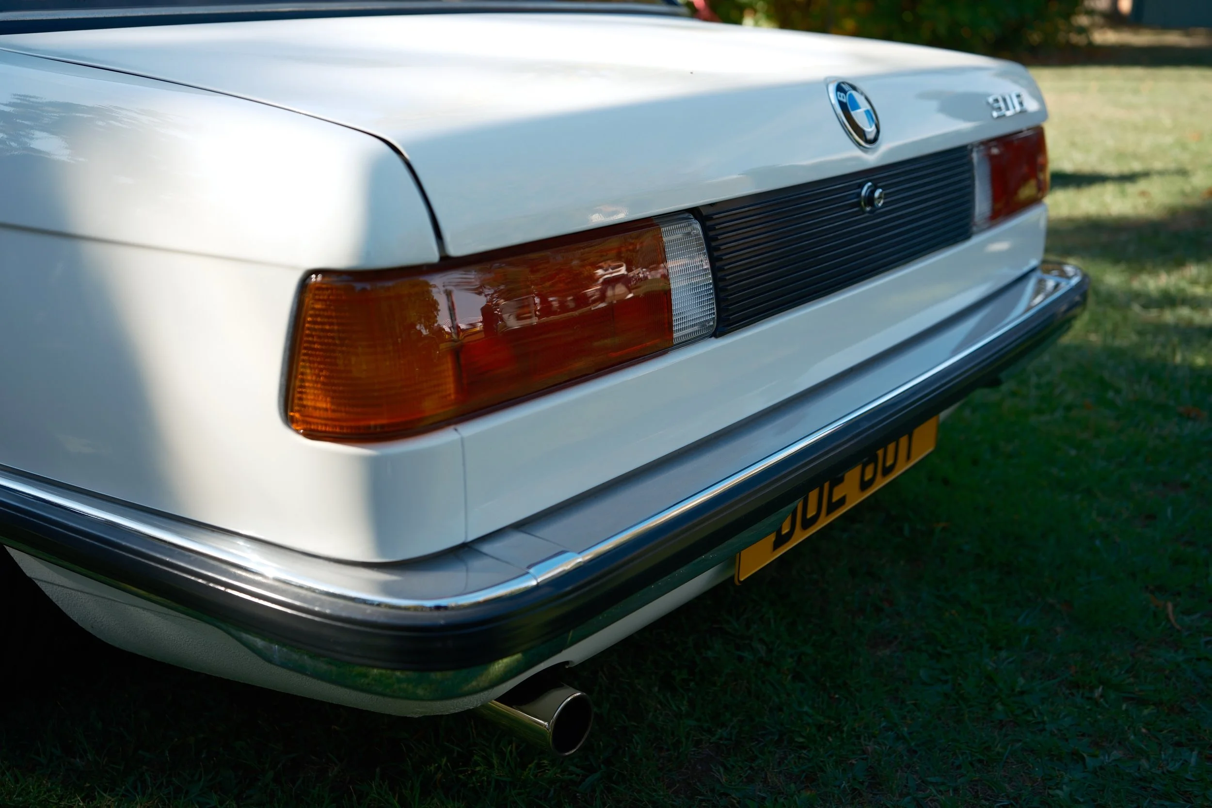 Rear view of a white BMW 3 series car parked on grass, showing the taillights, rear emblem, and exhaust pipe.