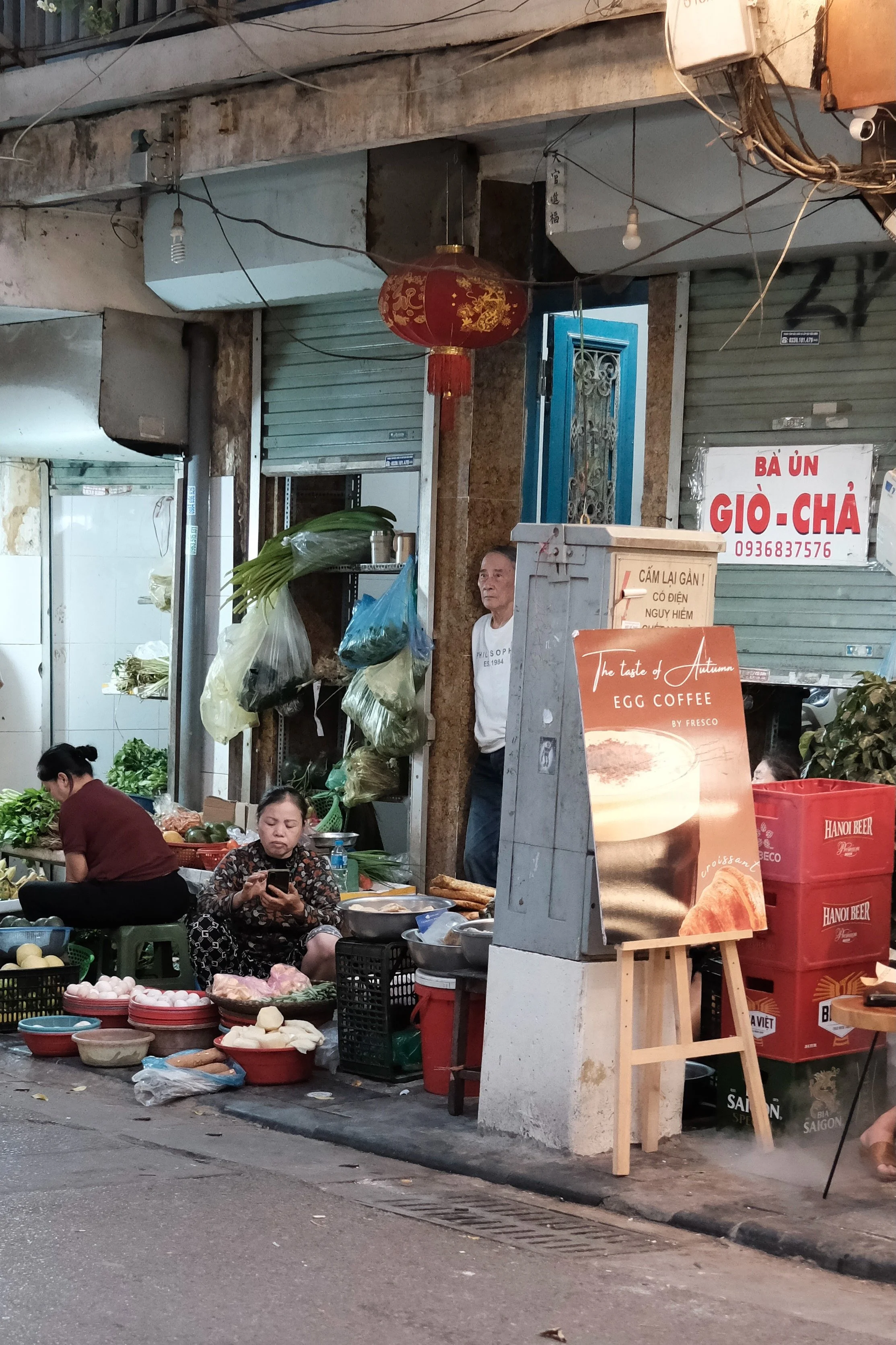 Street scene at a market stall in an Asian country, showing vendors selling vegetables and other produce, with some people sitting and using their phones.