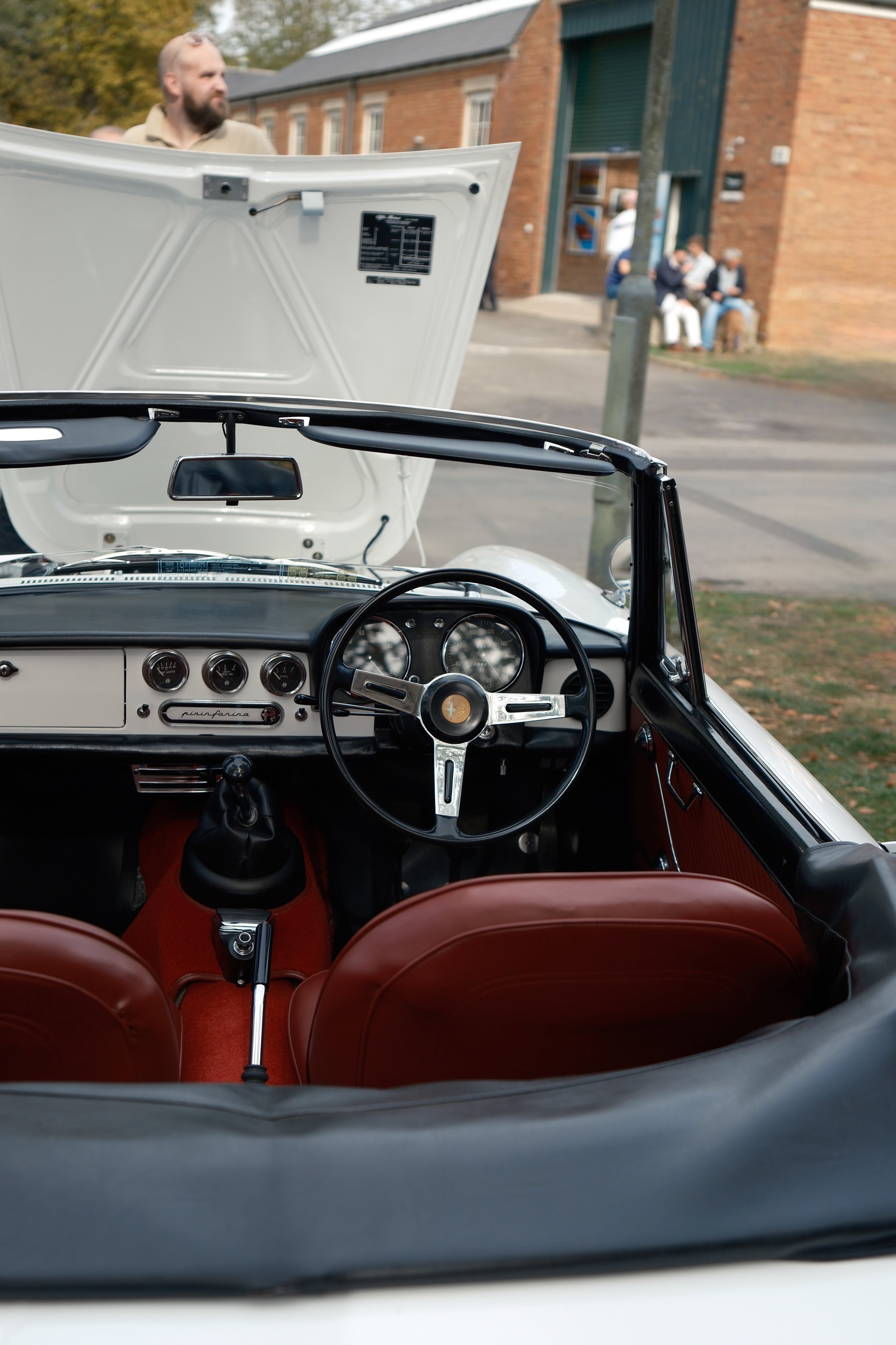 View from inside a vintage convertible car, showing the dashboard, steering wheel, and red leather seats, with the hood open and a person standing outside in a beige shirt, near a brick building with a group of people sitting on benches in the backgr