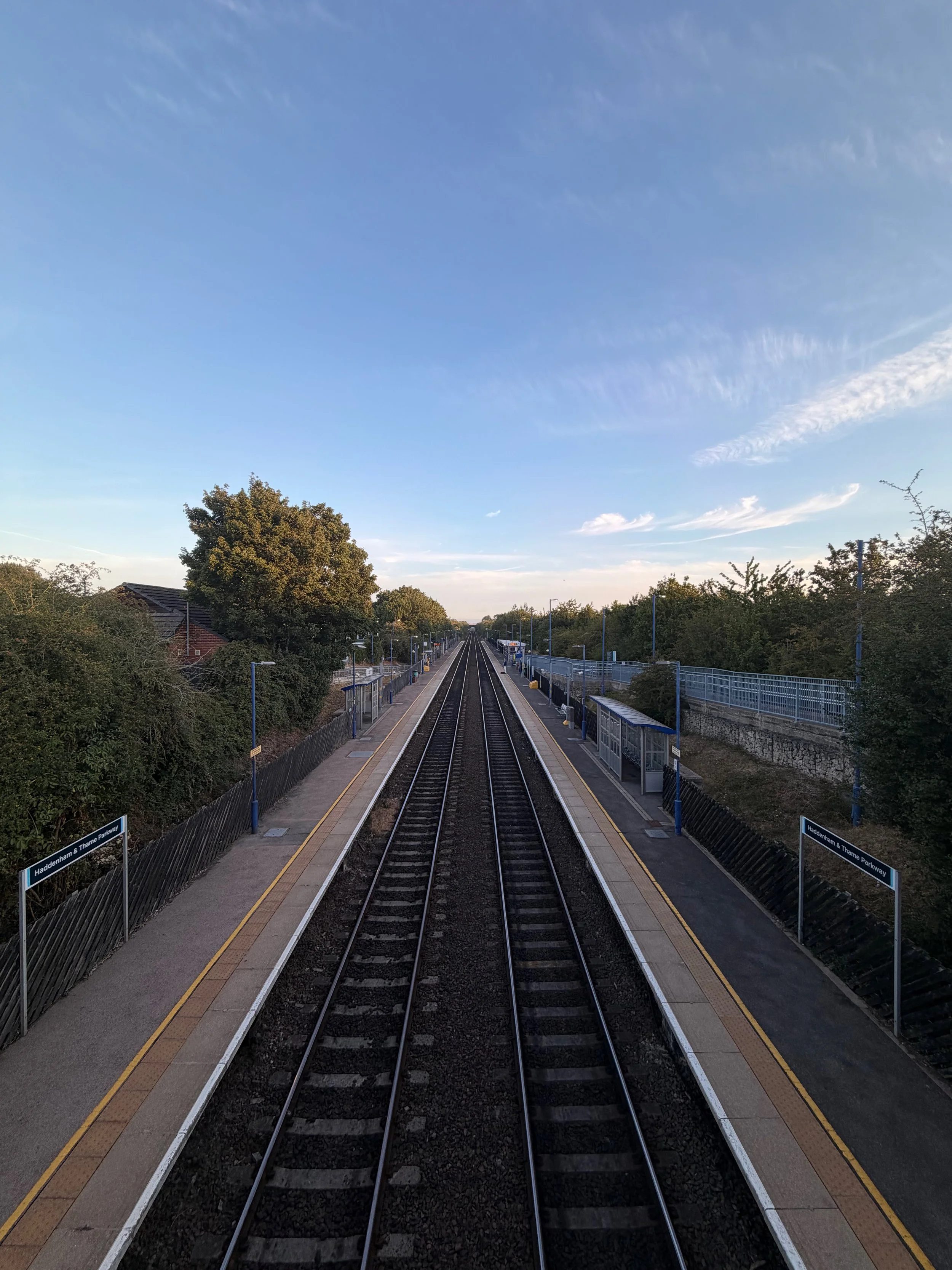 View of a train station platform with train tracks running through the center, surrounded by lush green trees, and a clear blue sky overhead.
