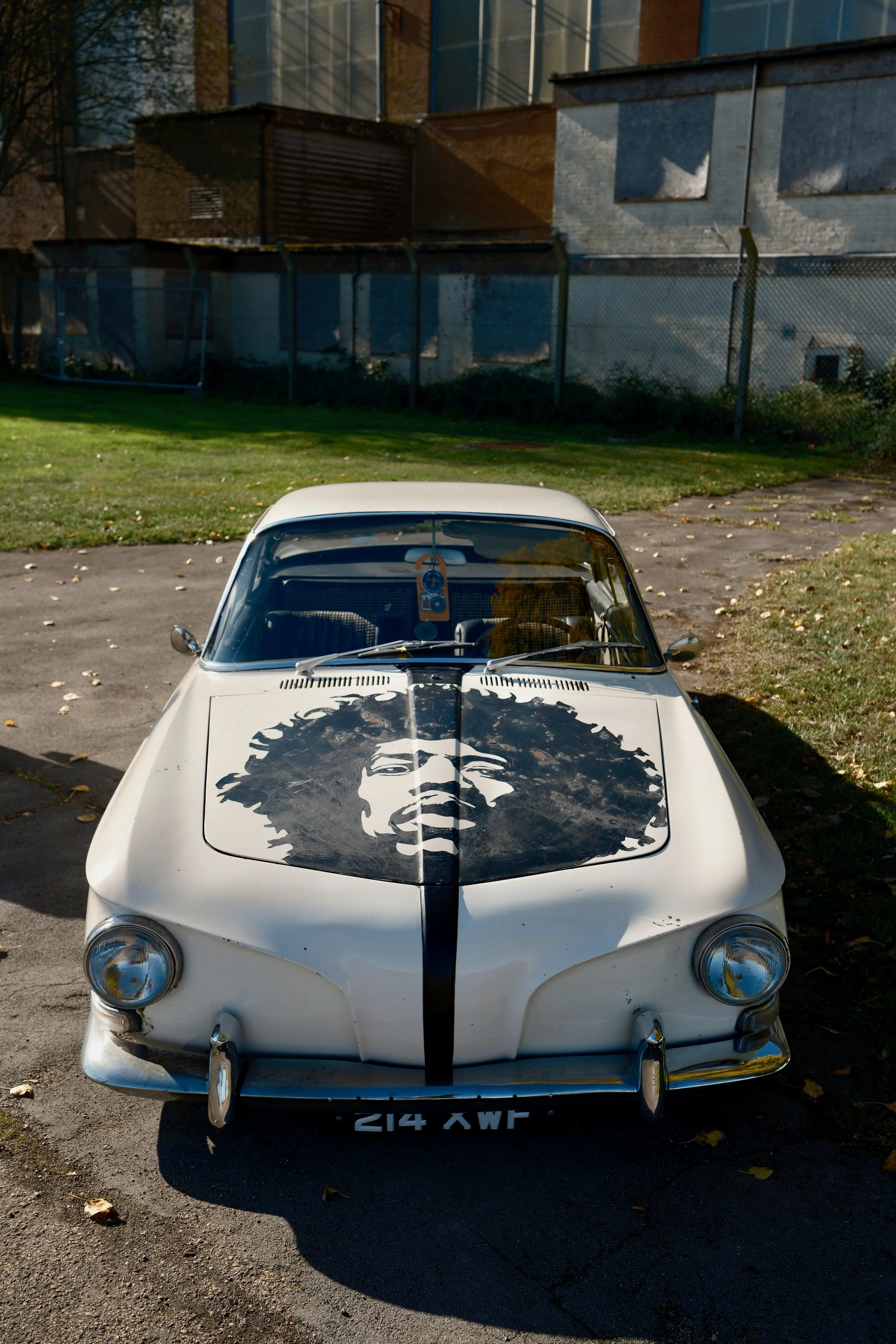 White vintage car with a large black and white portrait of a woman's face on the hood, parked outdoors on a paved area with fallen leaves, trees, a green grassy area, and a multi-story building in the background.