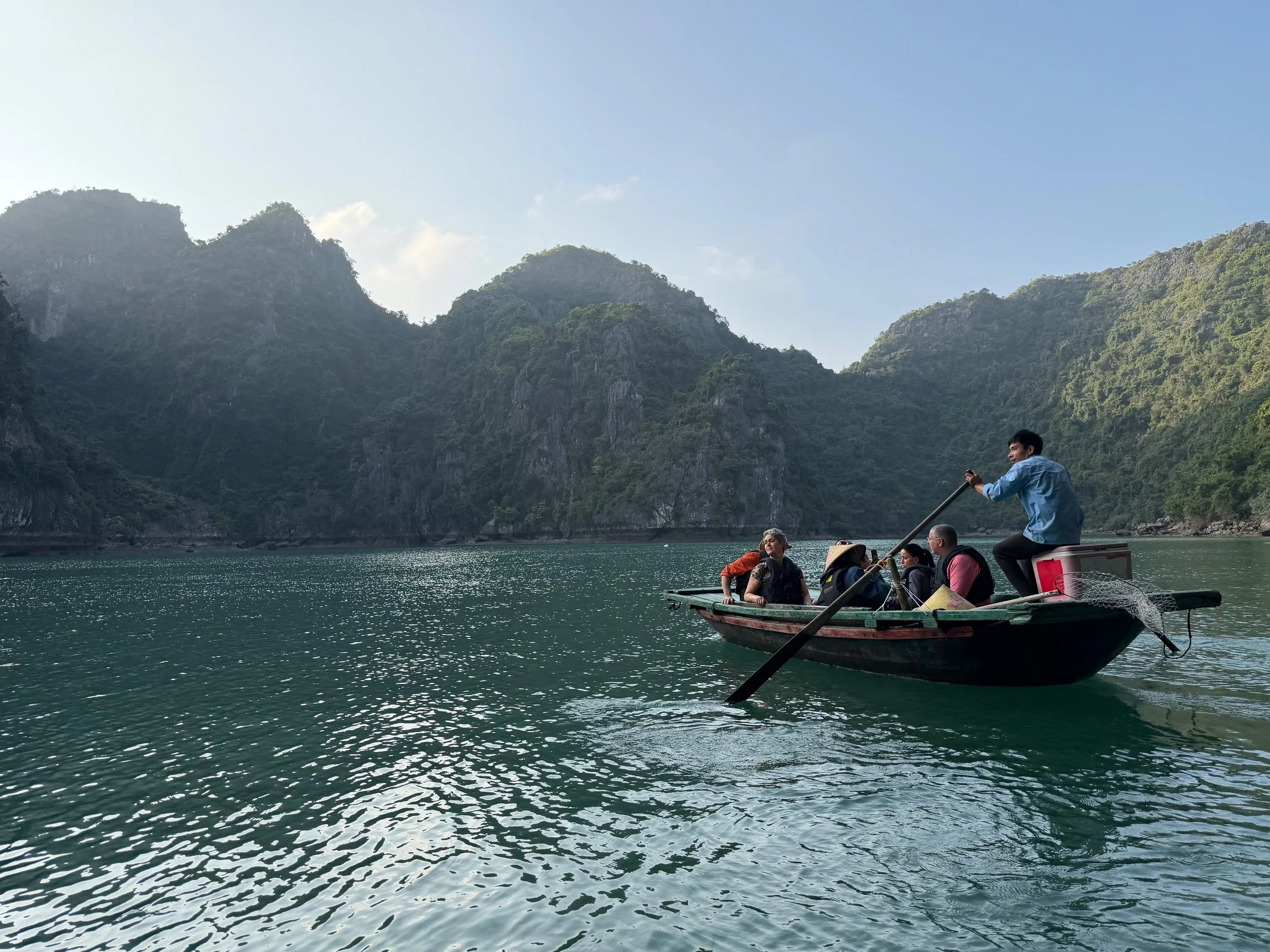 A group of people on a boat floating on a body of water surrounded by lush green mountains under a partly cloudy sky.