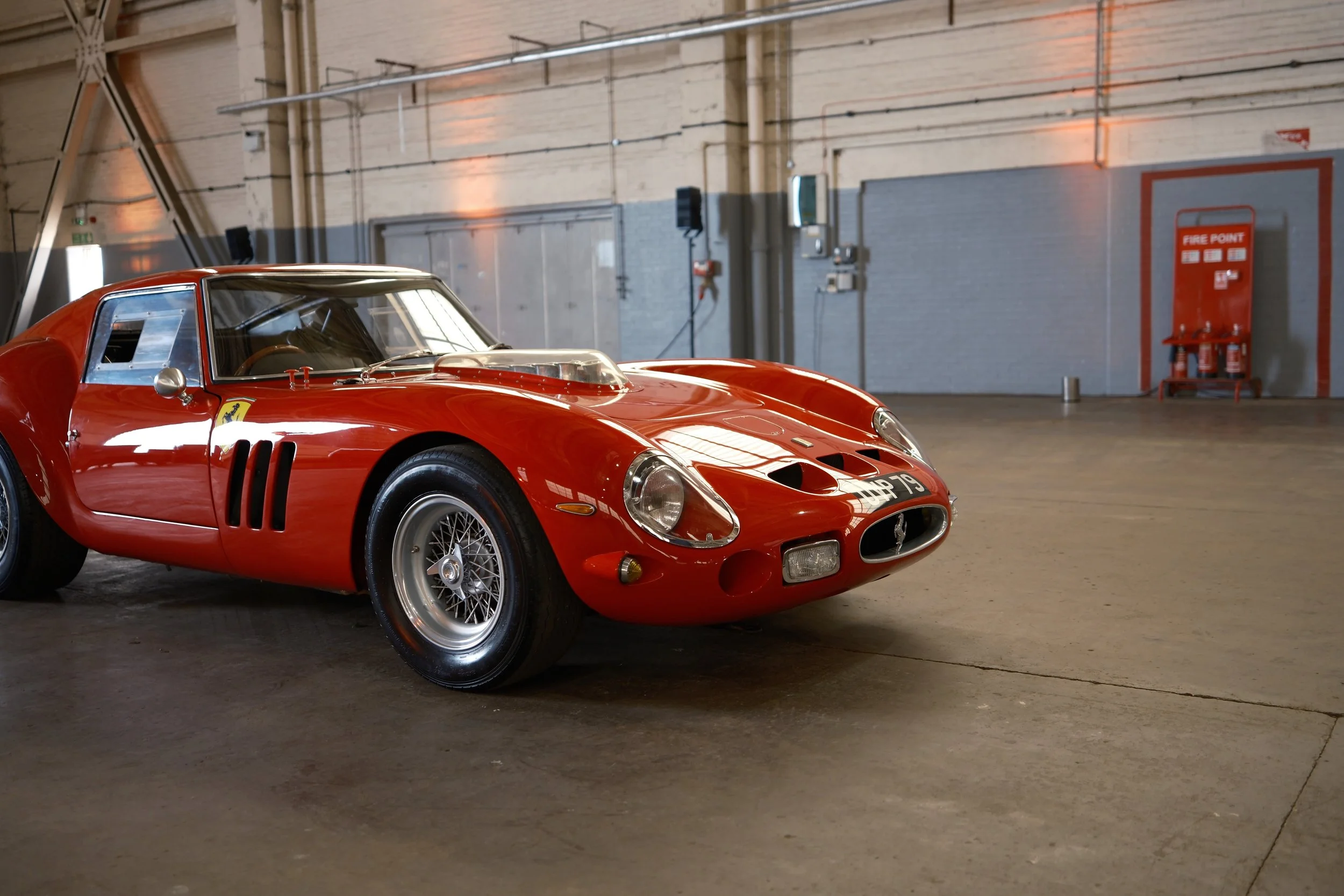 A vintage red Ferrari race car parked inside an industrial-looking garage with pipes and electrical panels on the wall, a fire extinguisher, and a fire point sign.