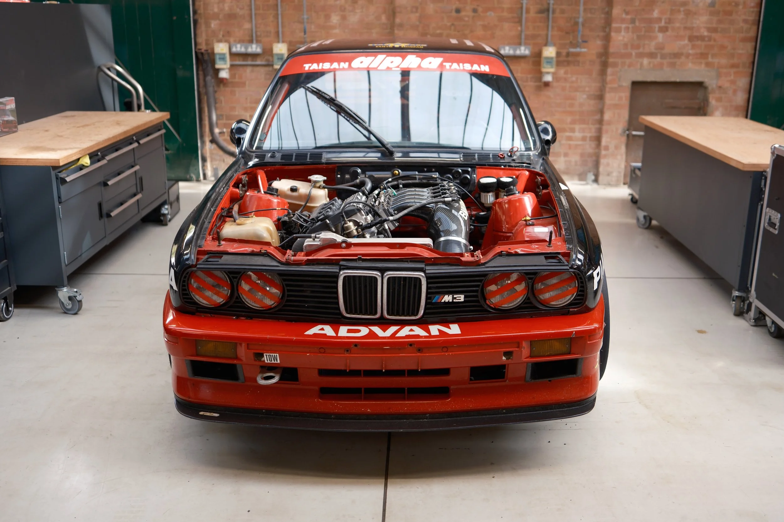 Front view of a red race car with its hood open, revealing the engine, in a workshop garage.