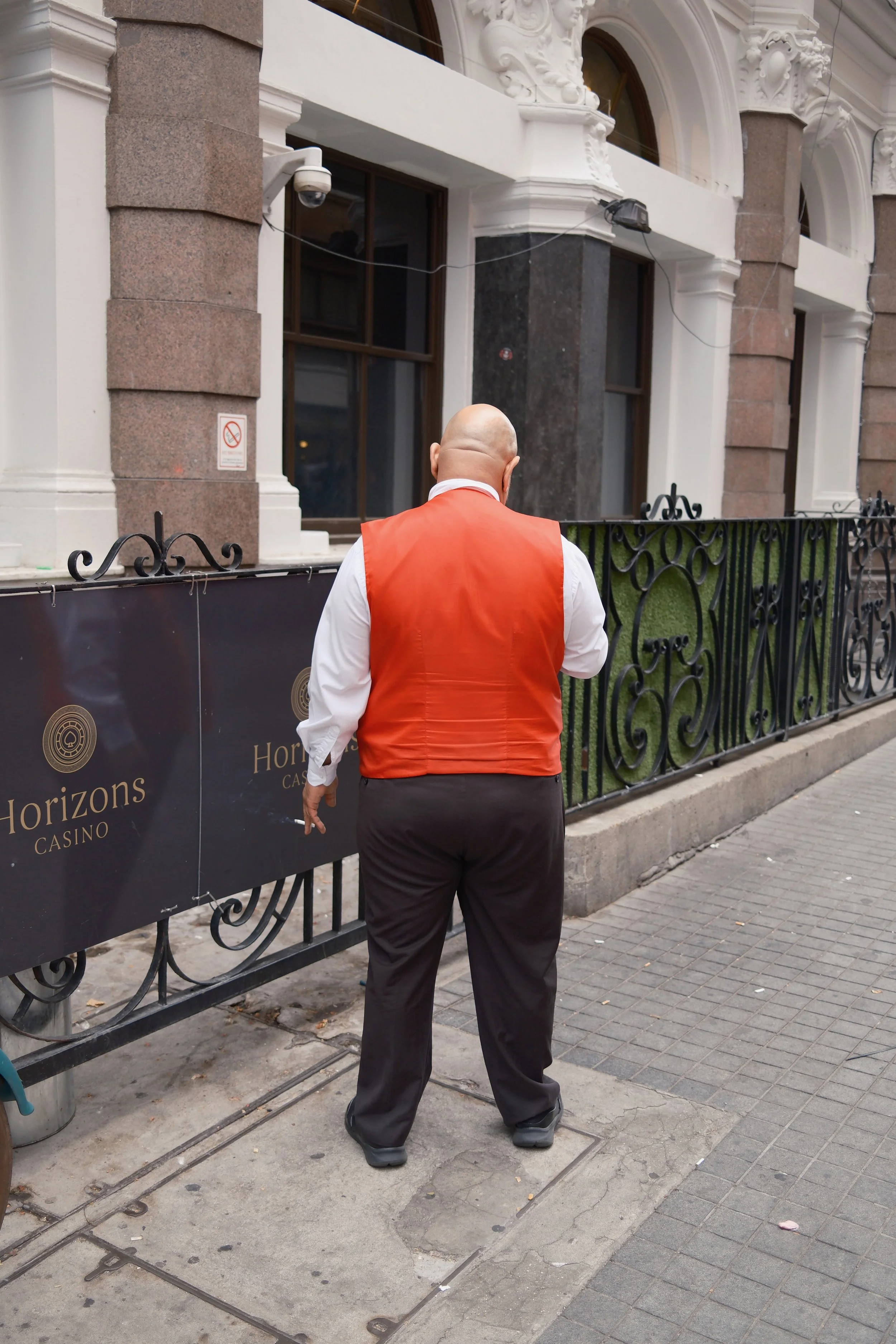 A person in a white shirt and orange vest stands on a city sidewalk, facing away from the camera, in front of a black wrought-iron fence and a building with ornate white architectural details.