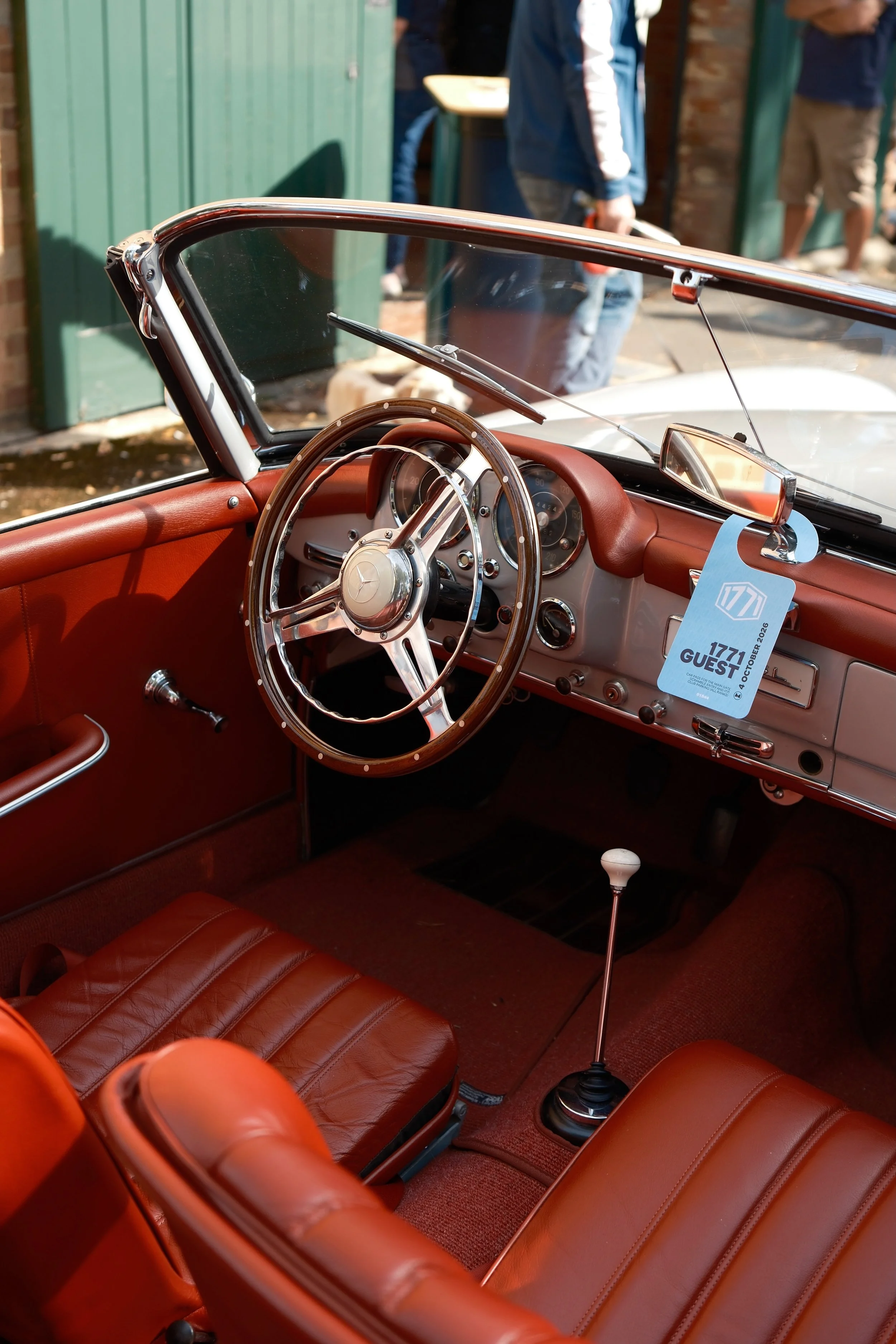 Interior of a vintage car with a brown leather dashboard and seats, a wooden steering wheel, and a gear shift. The car has a convertible top, and a parking permit hangs from the rearview mirror. People are visible outside, near a green building.
