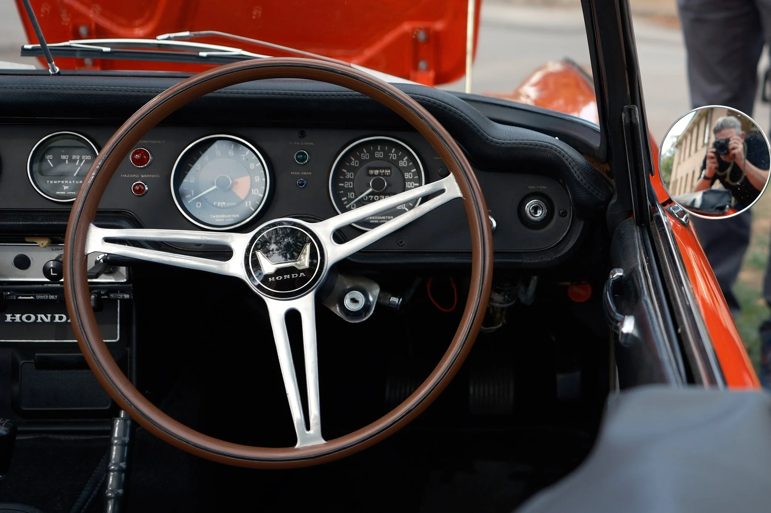 Close-up view of the dashboard and steering wheel of a vintage Honda car, with the photographer's reflection visible in the side mirror.
