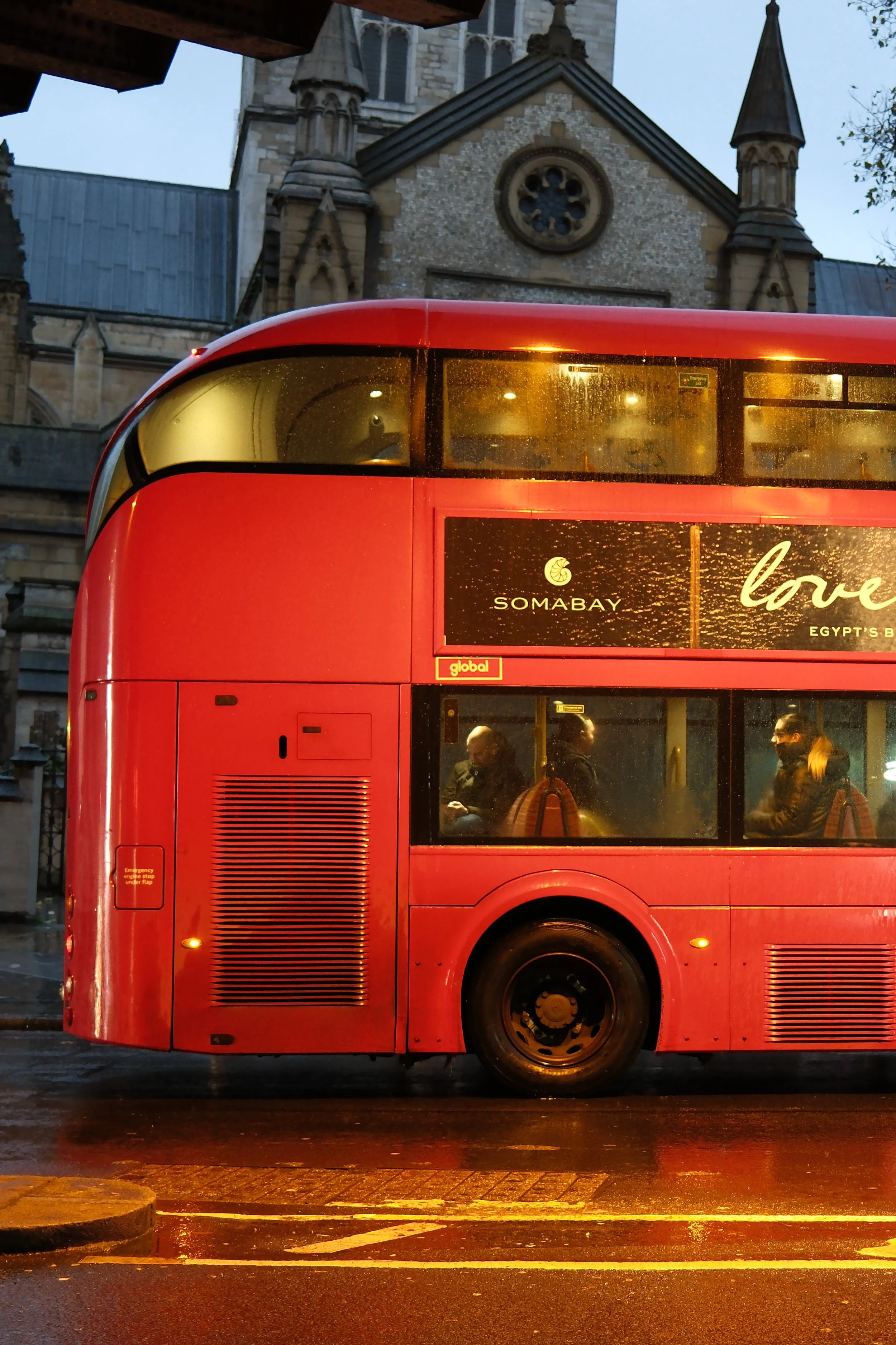 Red double-decker bus with digital advertisement on side, in front of historic stone church with gothic architecture and clock window, during evening rain.