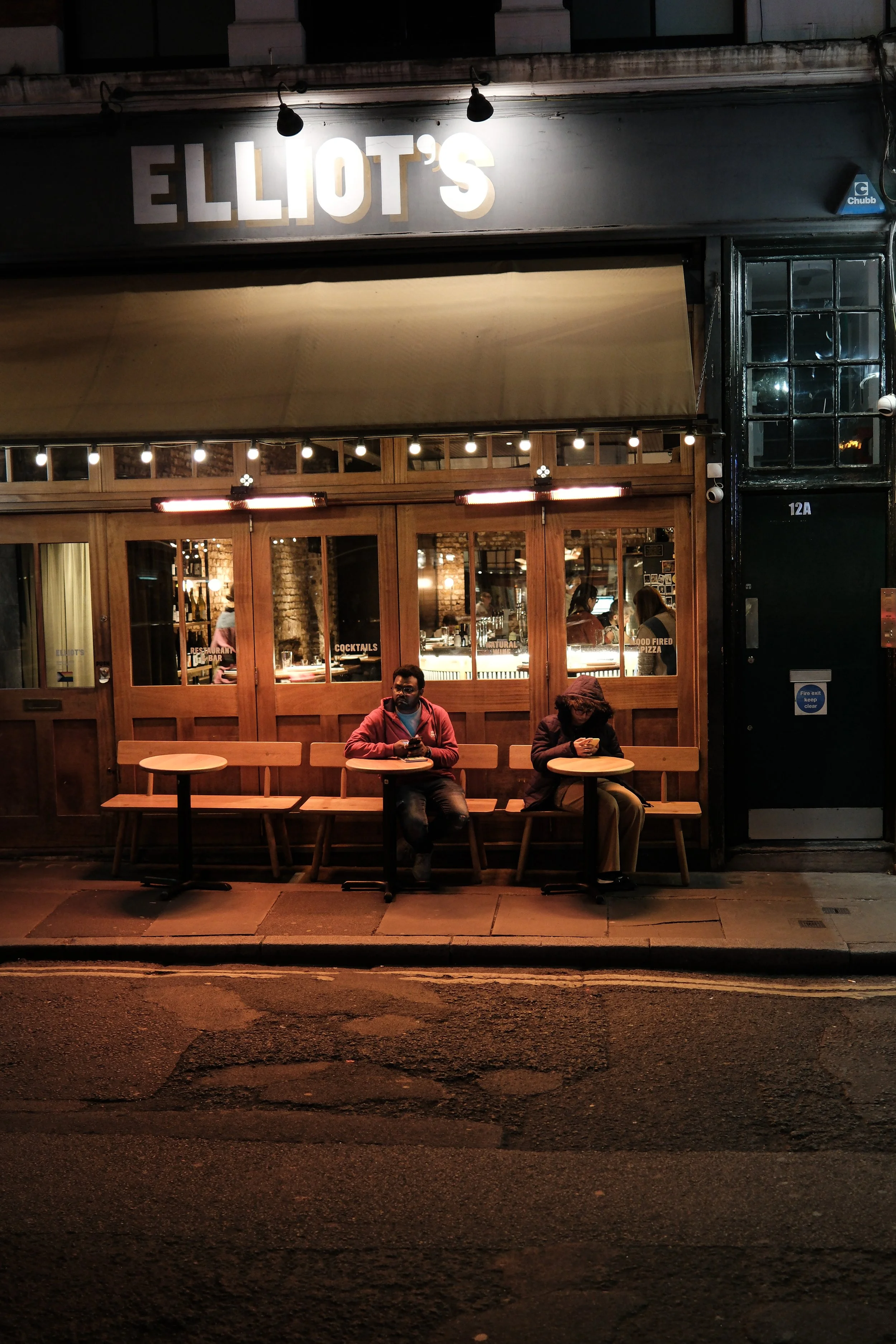 Night view of Elliot's restaurant with a dark exterior, illuminated sign, and large window showing the interior where people are seated, some using their phones, and warm lighting.