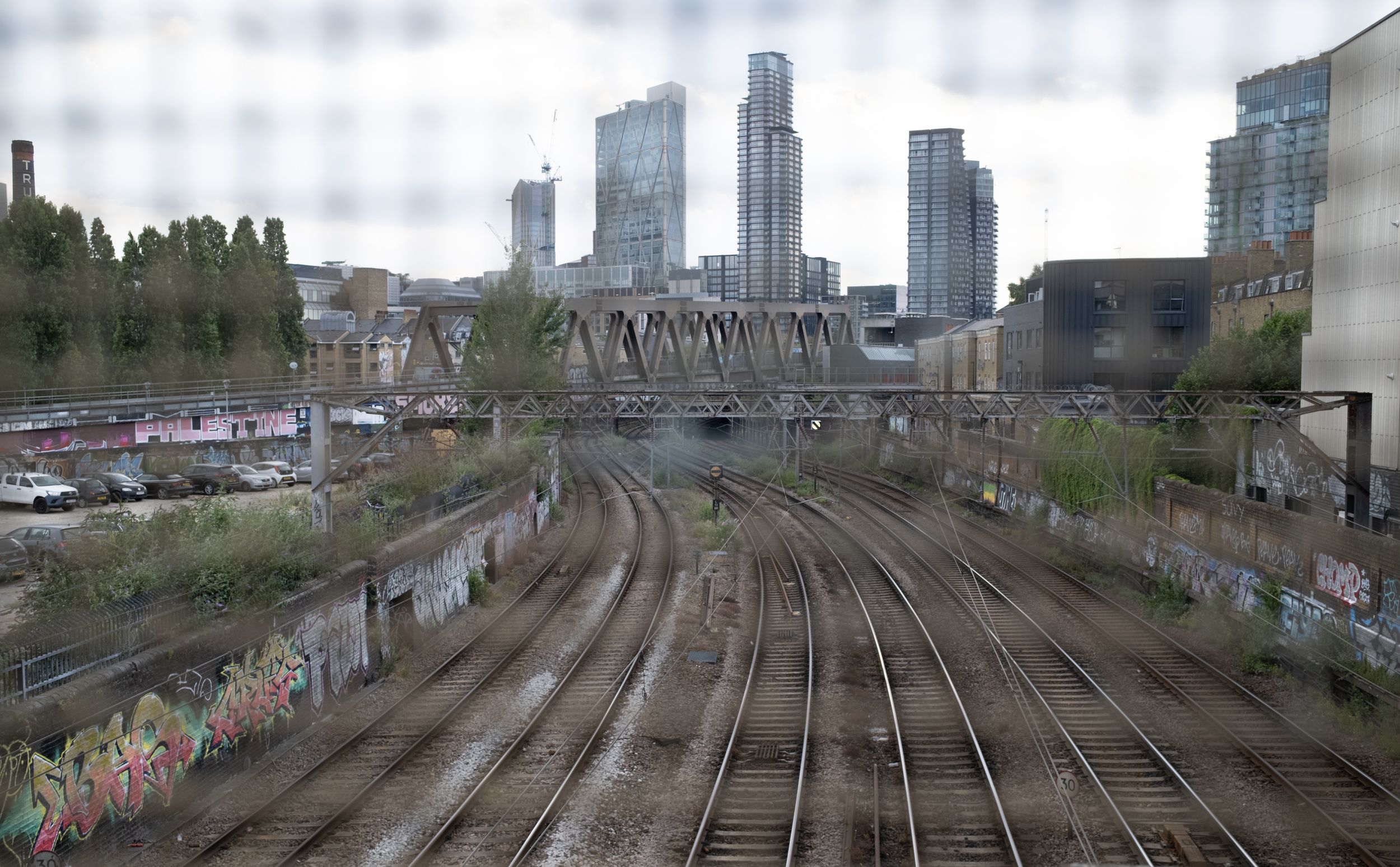 City skyline with tall modern buildings, railway tracks, and graffiti-covered walls in the foreground. Overcast sky.