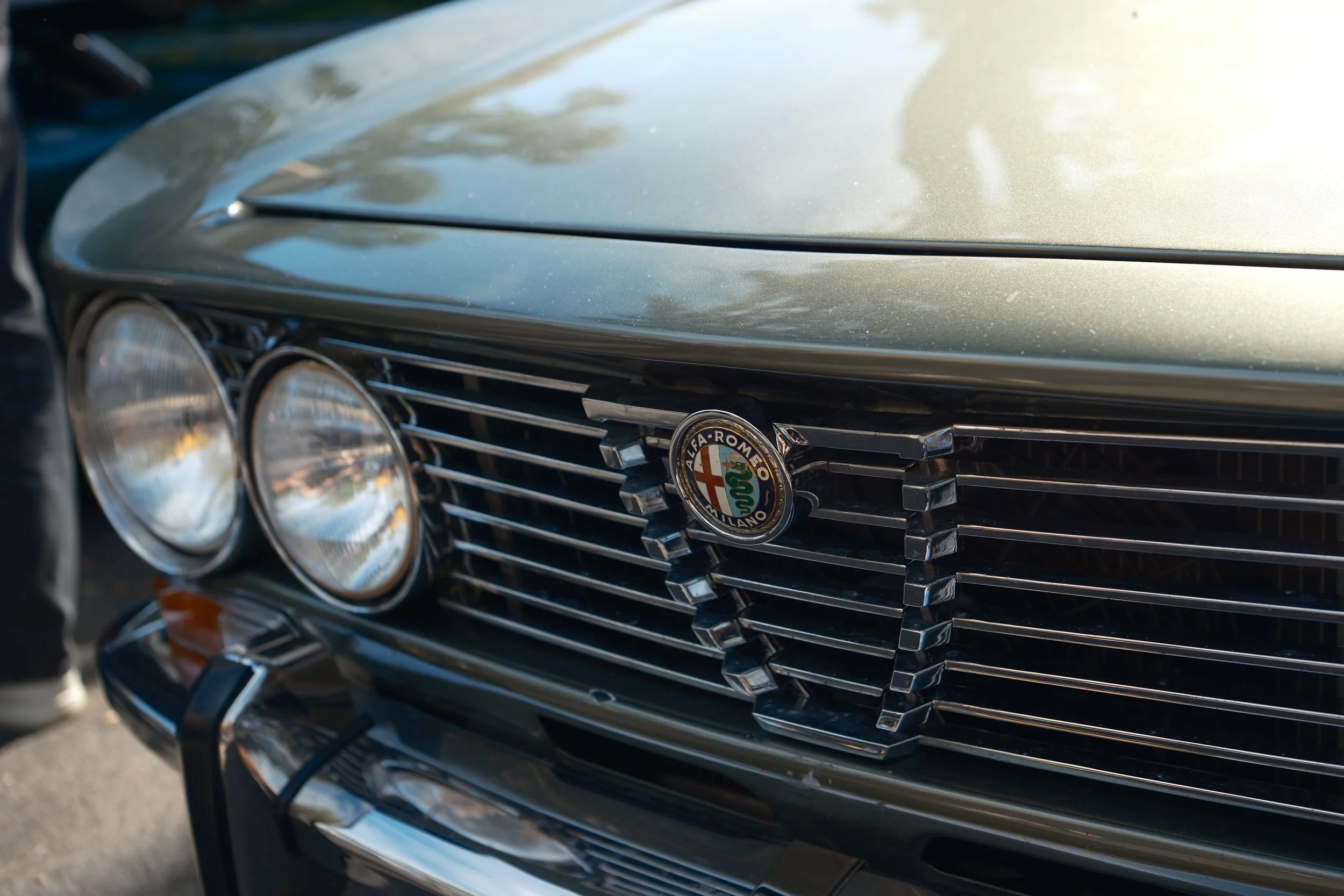 Close-up of the front grille of a vintage Alfa Romeo car, featuring chrome accents and the Alfa Romeo emblem, with a reflective metallic green hood.