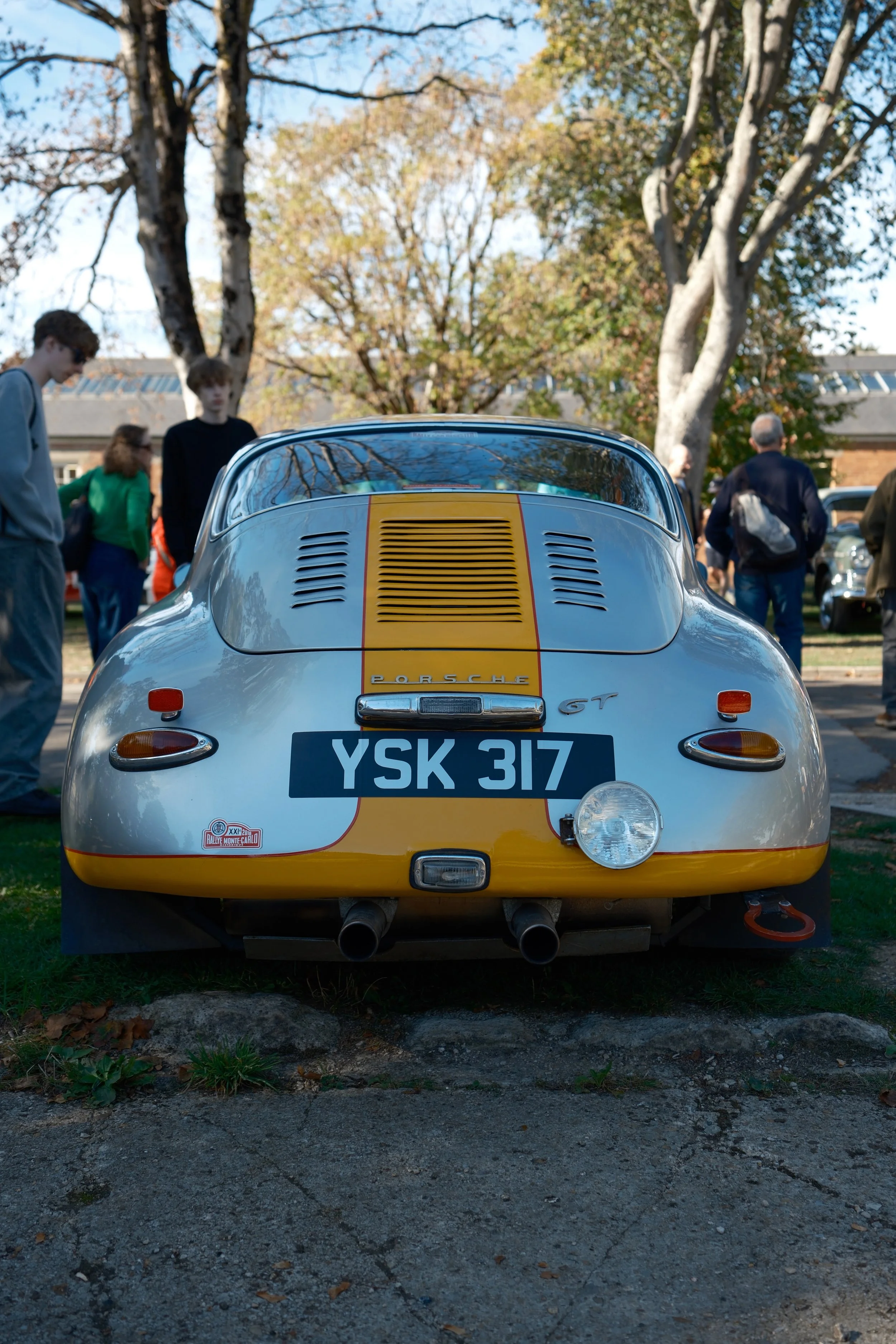 Rear view of a classic Porsche 356 GT sports car with a white and yellow paint job, parked outdoors on a gravel surface, with people and trees in the background.