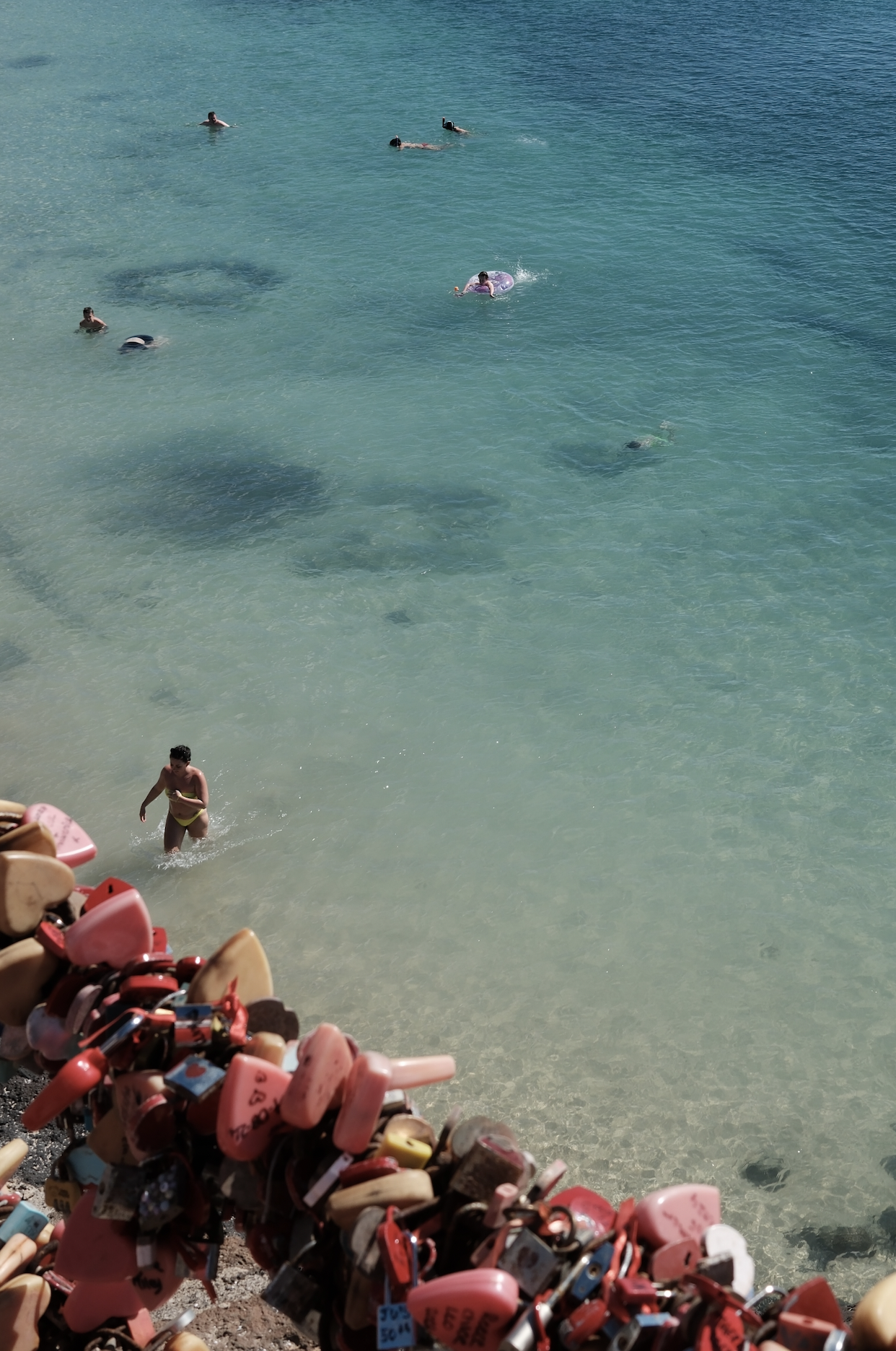 Many people swimming and floating in the ocean near a sandy beach. In the foreground, a pile of love locks attached to a rope or barrier.