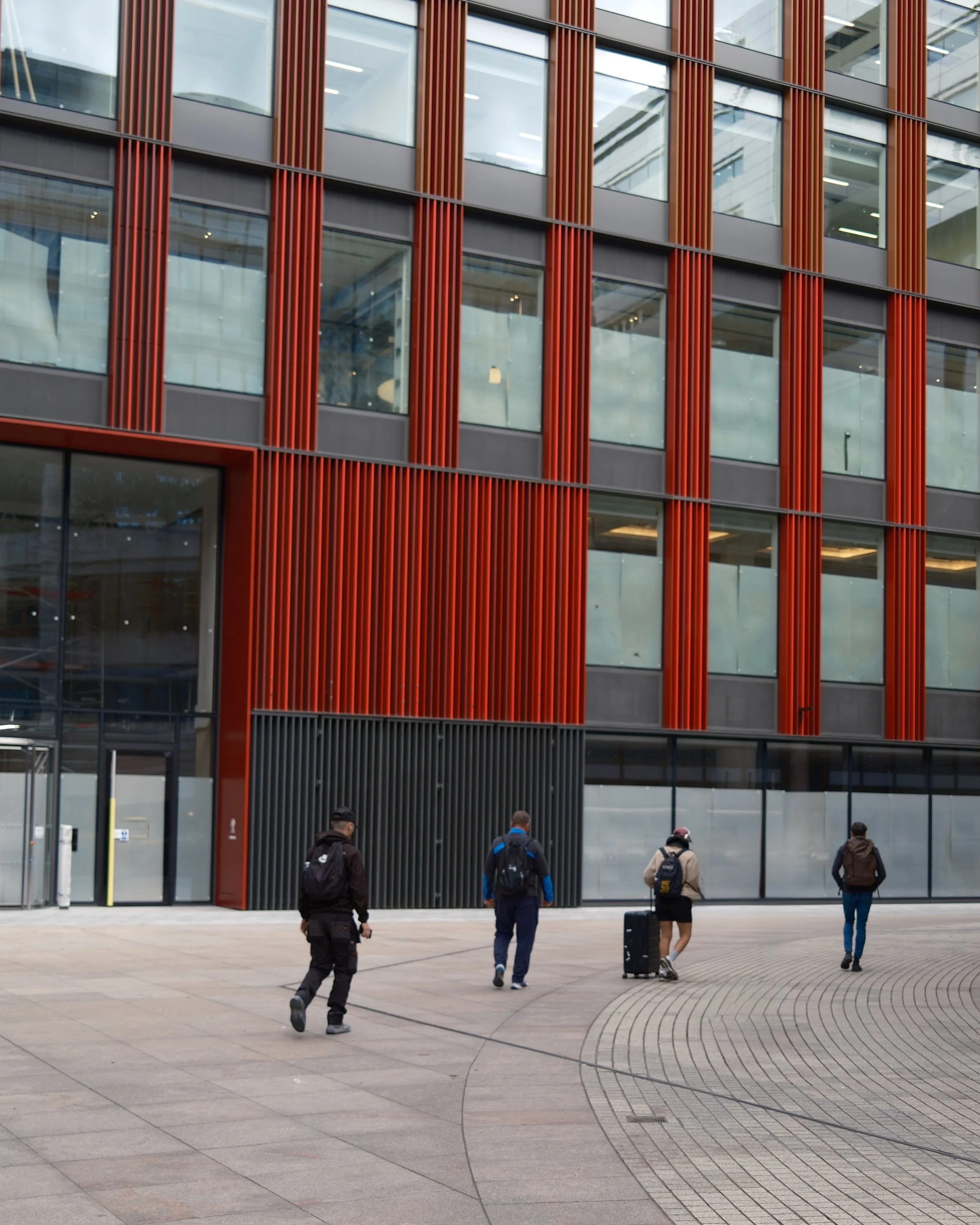 People walking outside a modern glass building with red vertical metal accents.