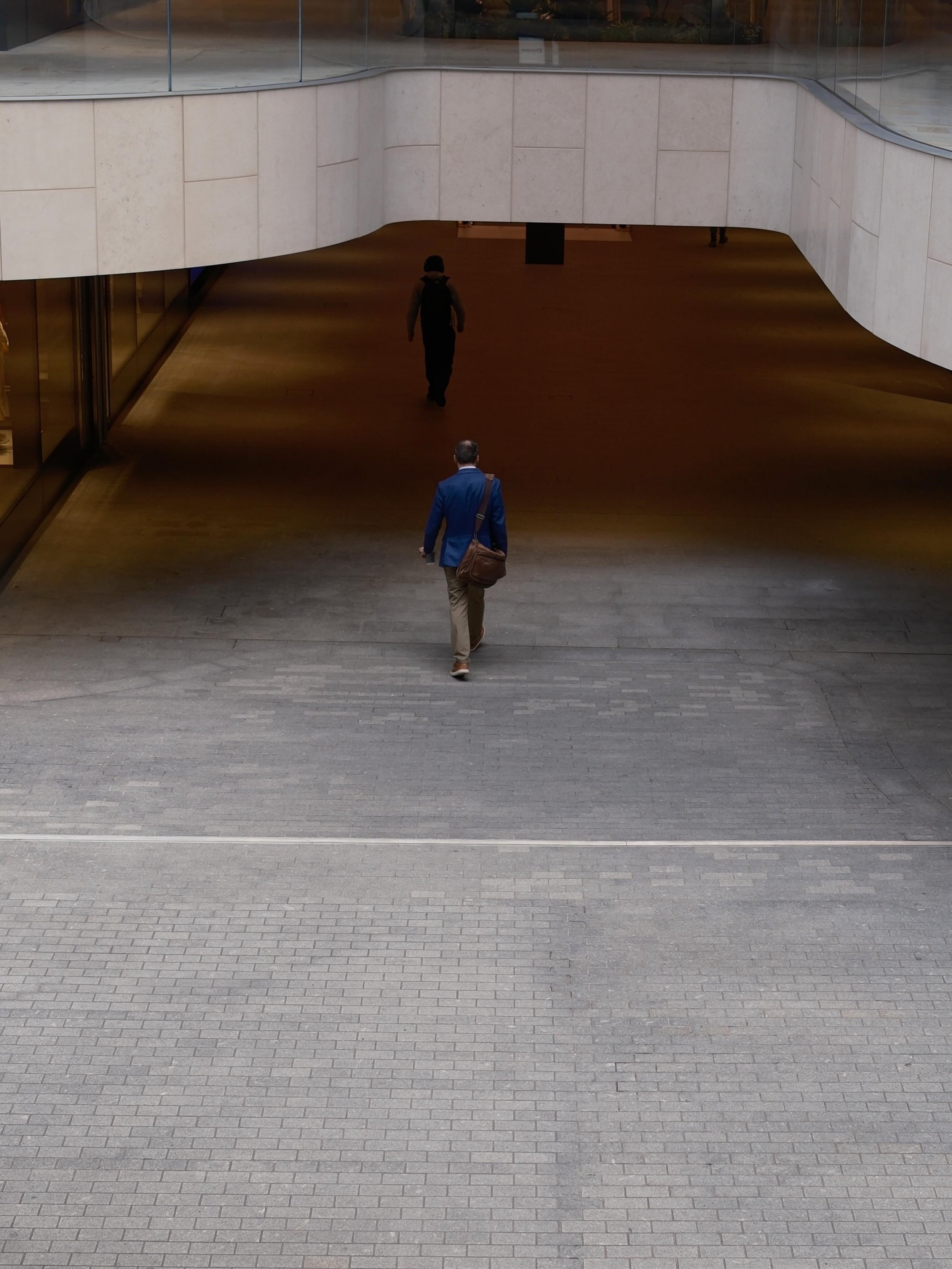 Two people walking in an indoor mall or building with modern architecture. The person in the foreground is wearing a blue suit with a brown shoulder bag, and the person in the background is dressed in dark clothing with a backpack. The floor is made 