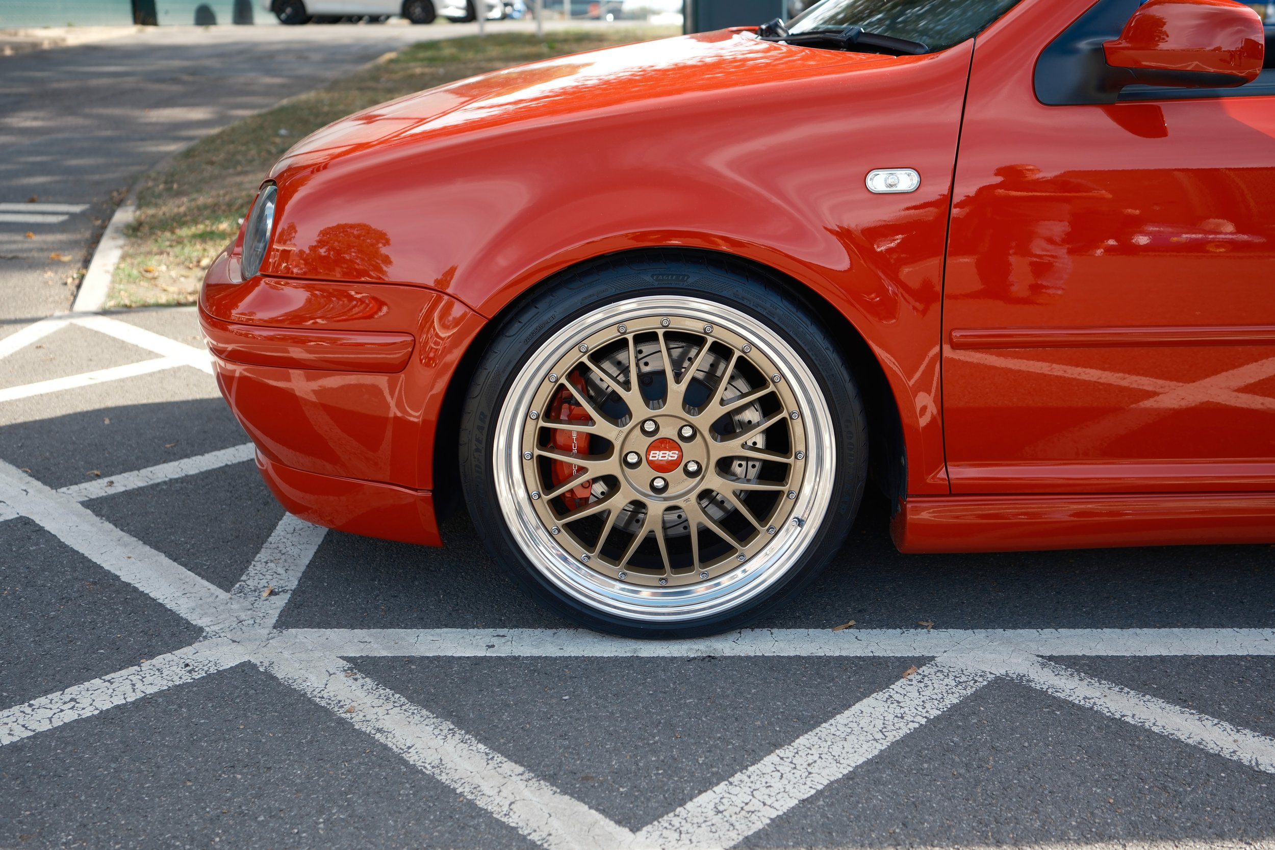Close-up of a red sports car parked in a parking lot with a multi-spoke alloy wheel and a red brake caliper visible.