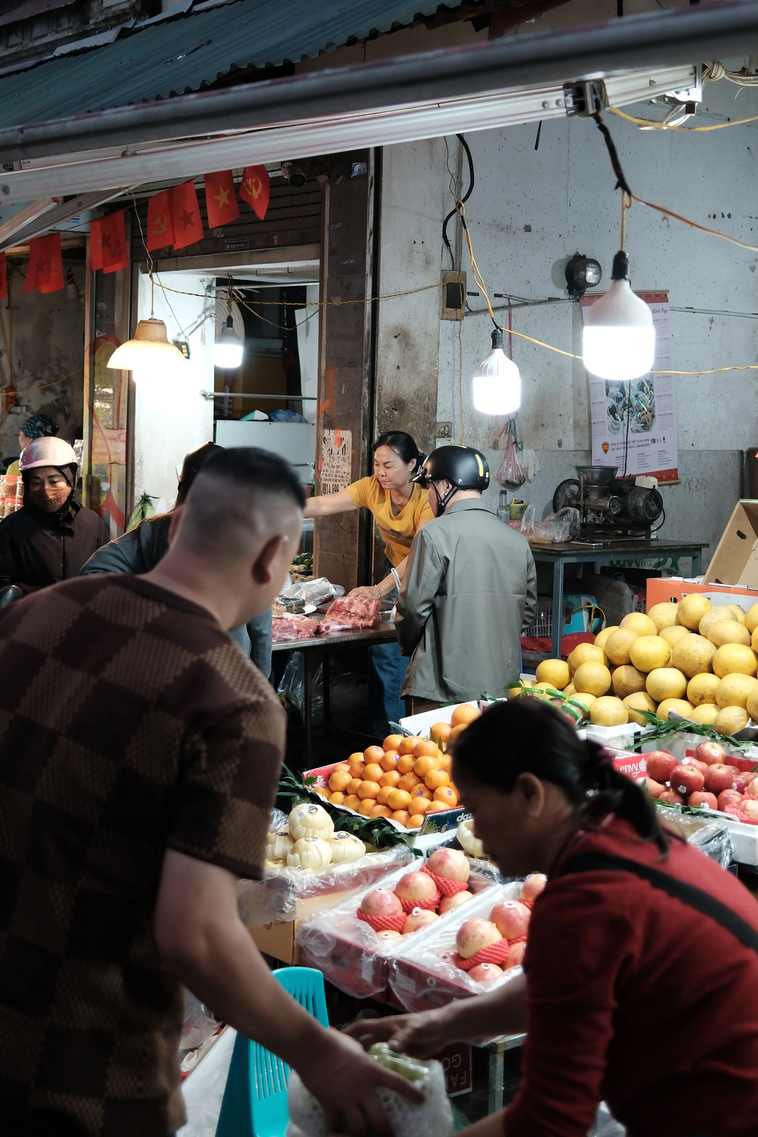 A busy marketplace with vendors selling fresh fruits like apples, oranges, and pomelos. People are examining and buying produce, and the background shows an old wall with hanging flags featuring communist symbols. Bright lights illuminate the scene.