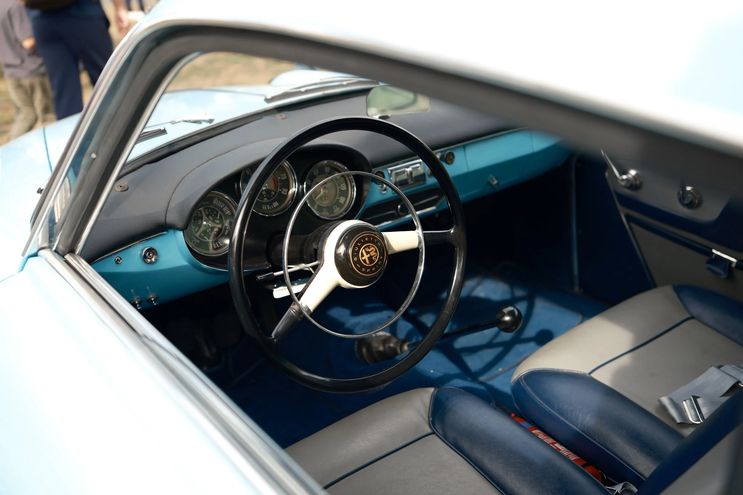 Interior of a vintage light blue car showing the dashboard with gauges and a steering wheel with an Alfa Romeo logo, and part of the passenger seat.