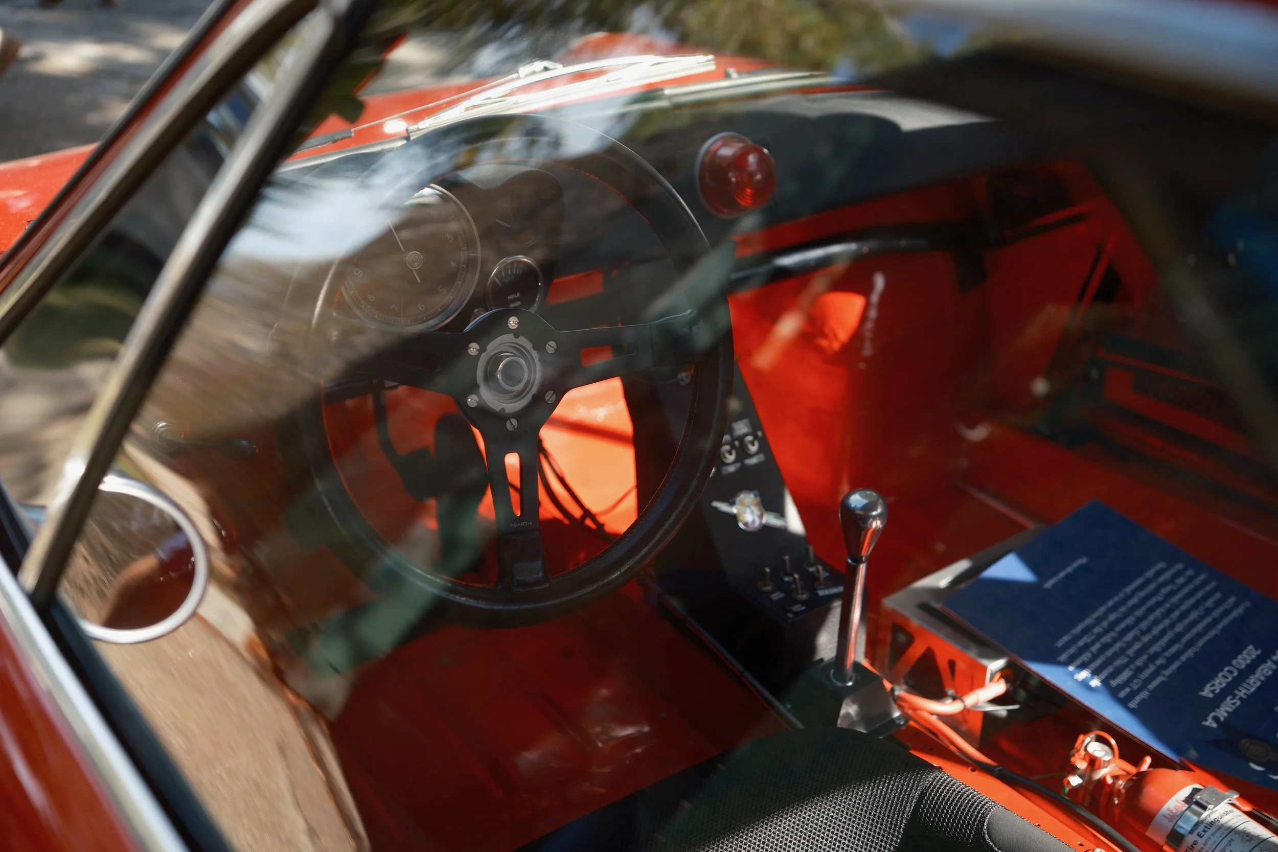 The interior of a vintage red race car showing a steering wheel, dashboard, gear shift, and a fire extinguisher, with reflections visible on the windshield.